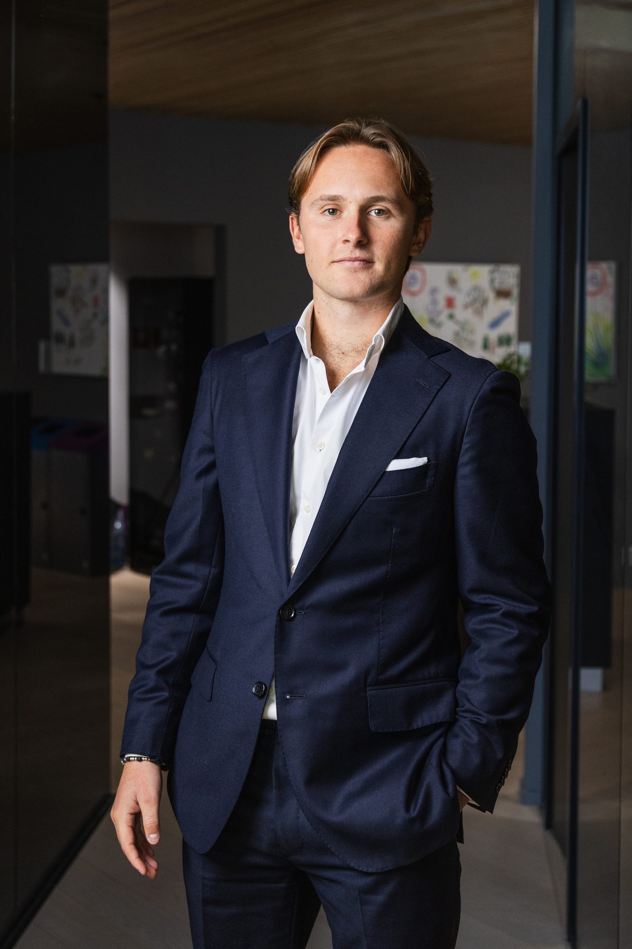 Man in navy suit and white shirt, standing with hand in pocket, looking at camera.