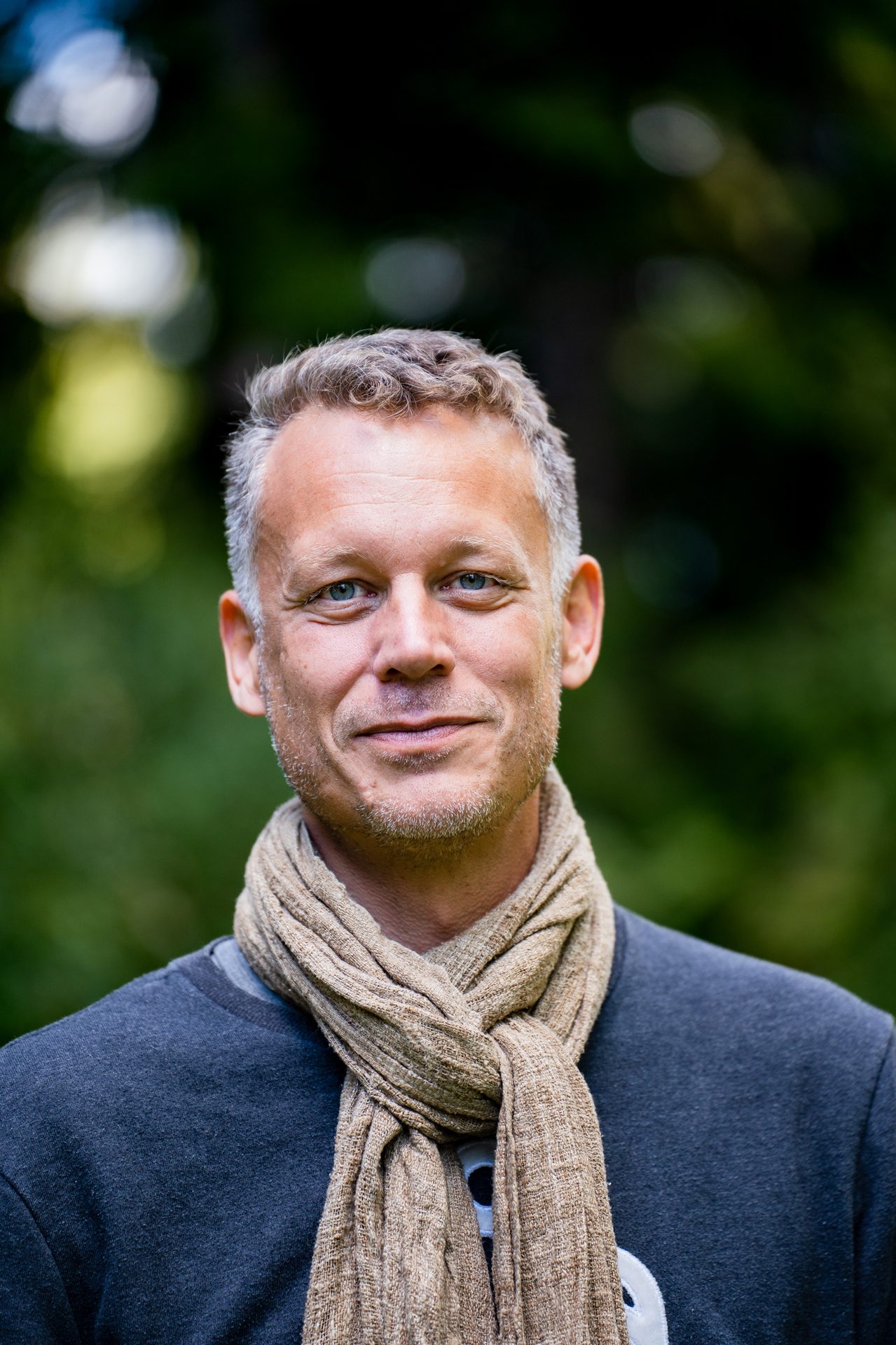 Smiling man with gray hair, blue eyes, dark sweater, and beige scarf outdoors.