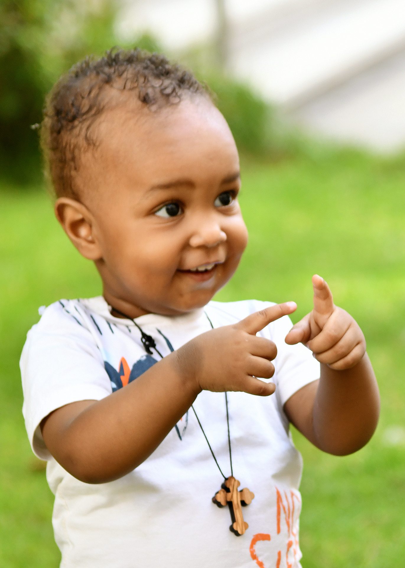 Smiling toddler with dark curly hair and a cross necklace, pointing fingers, green outdoor background.