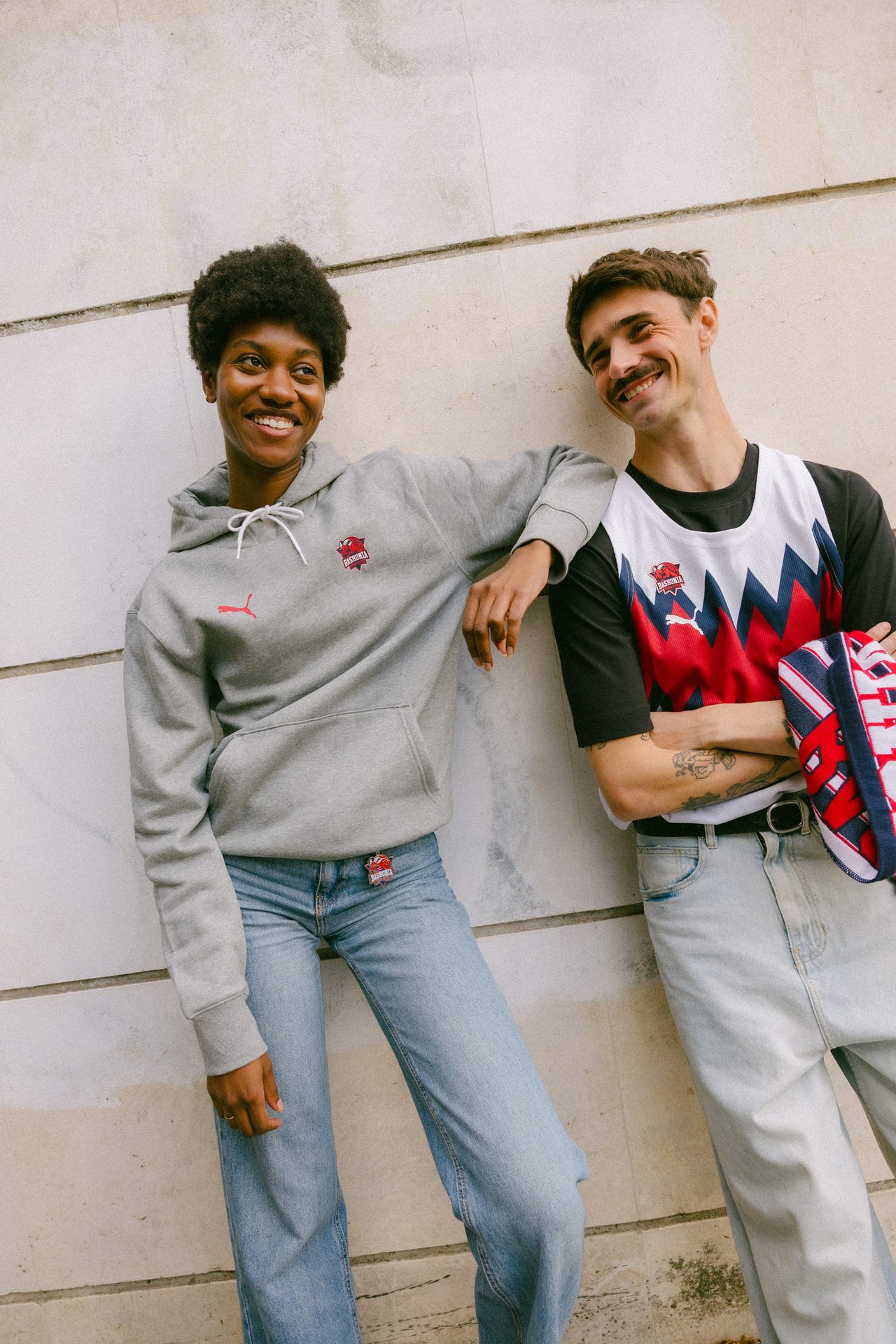 Smiling man and woman lean against a wall, wearing casual Baskonia-branded clothing.