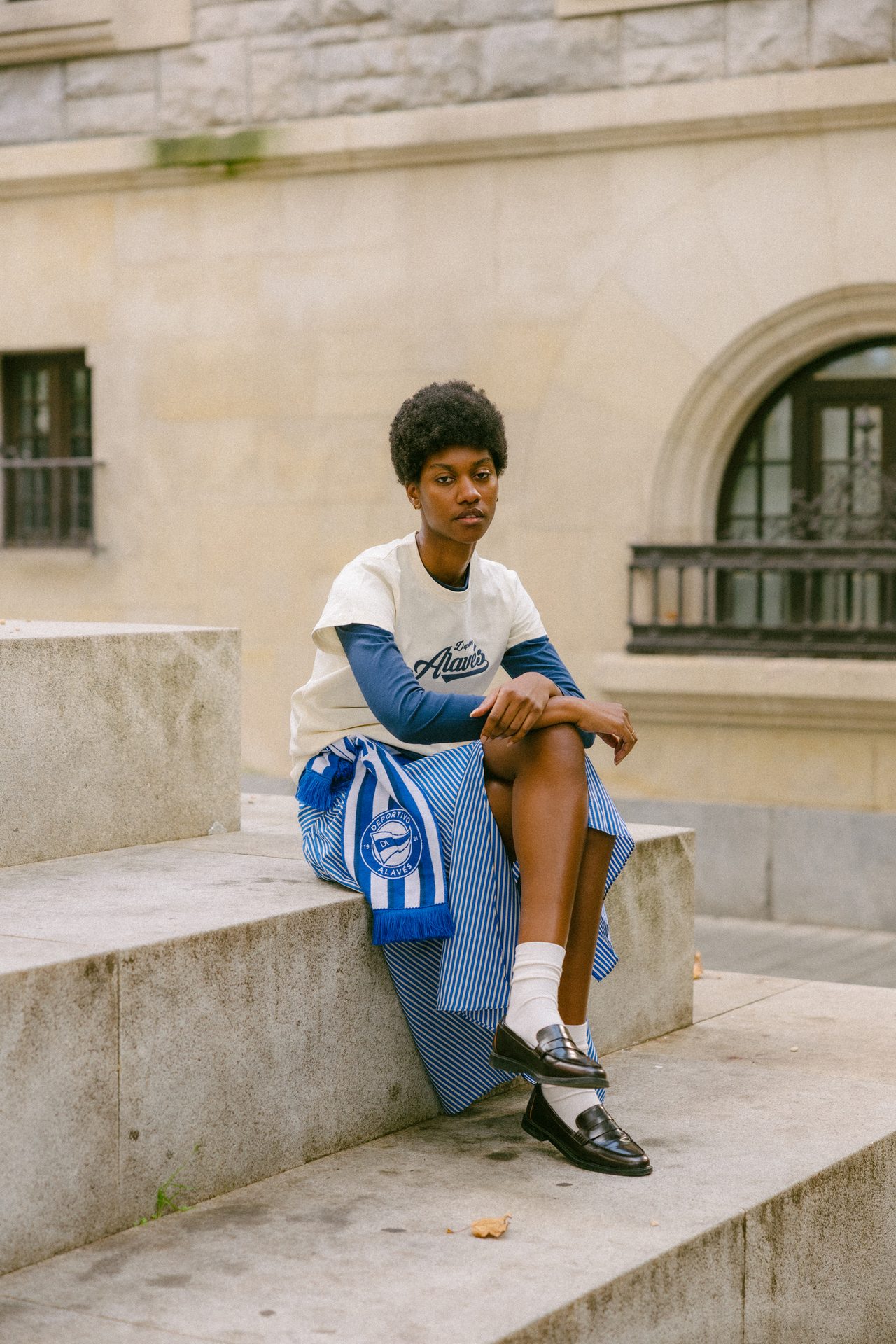 A person with an afro sits on steps in a layered top, striped skirt, loafers, and a blue/white scarf.