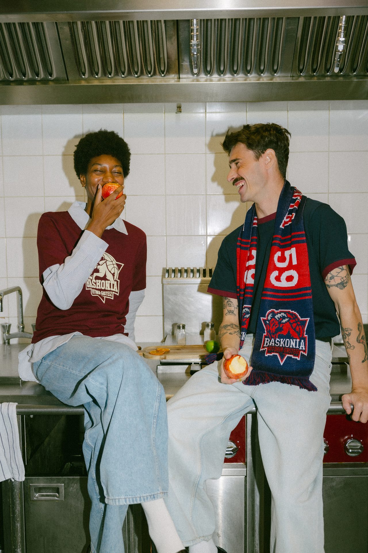 Two people in a kitchen, smiling and eating apples, wearing basketball fan gear.