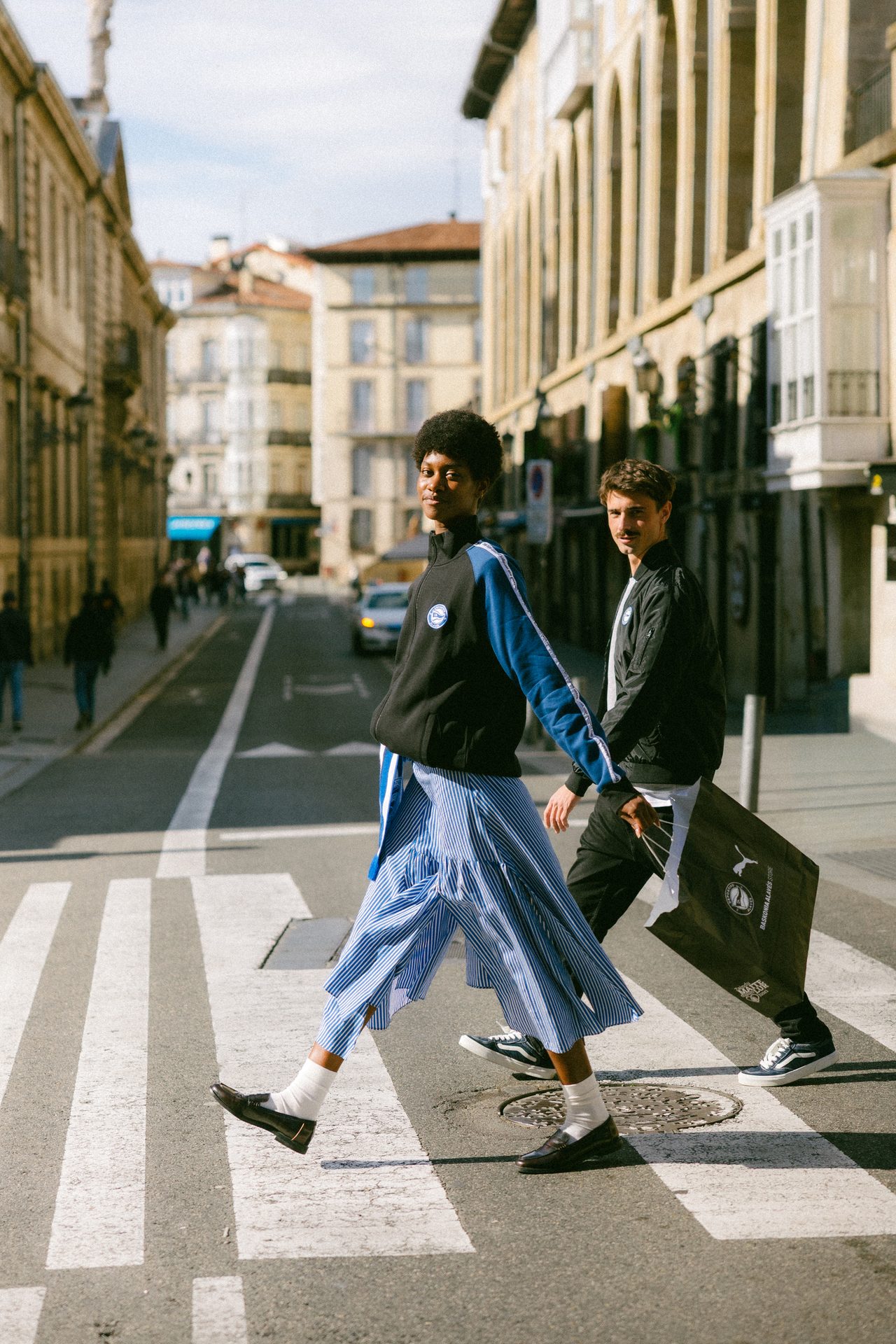 Two people in streetwear, a woman in a striped skirt and man with a bag, cross a street.