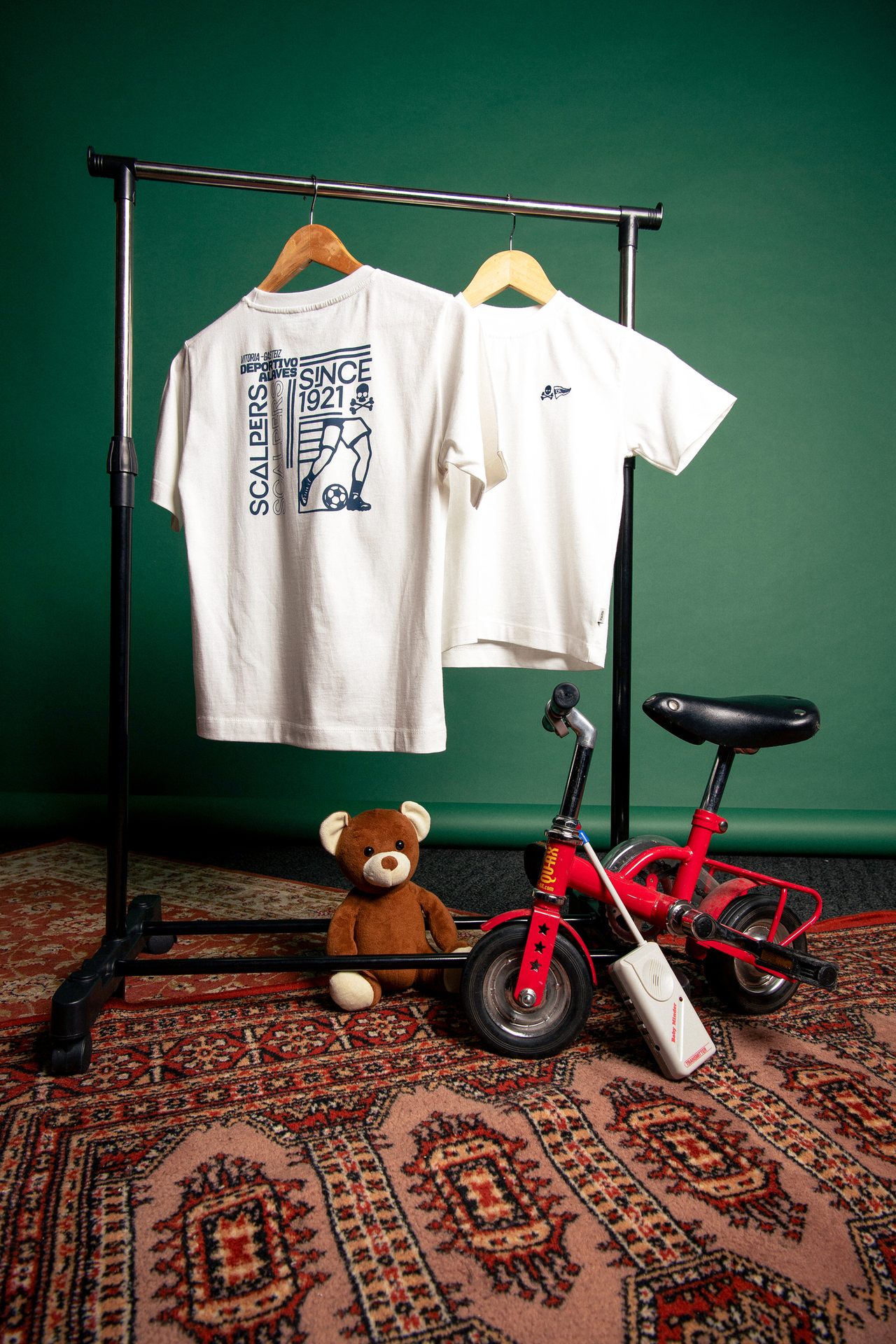 Two white t-shirts, a teddy bear, and a red toy tricycle on a rug against a green background.