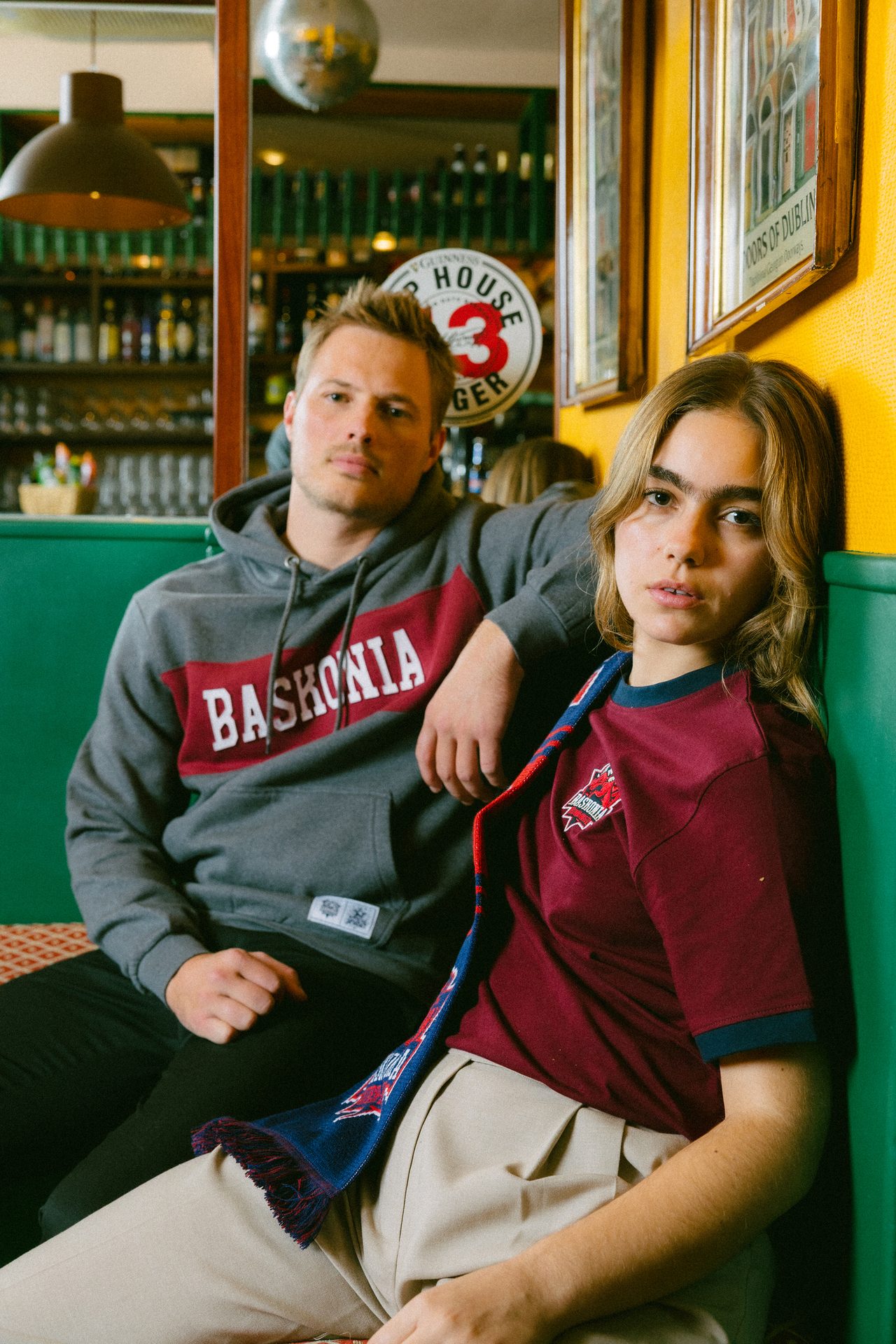 Man in "BASKONIA" hoodie and woman in maroon shirt with scarf sit in a colorful bar.