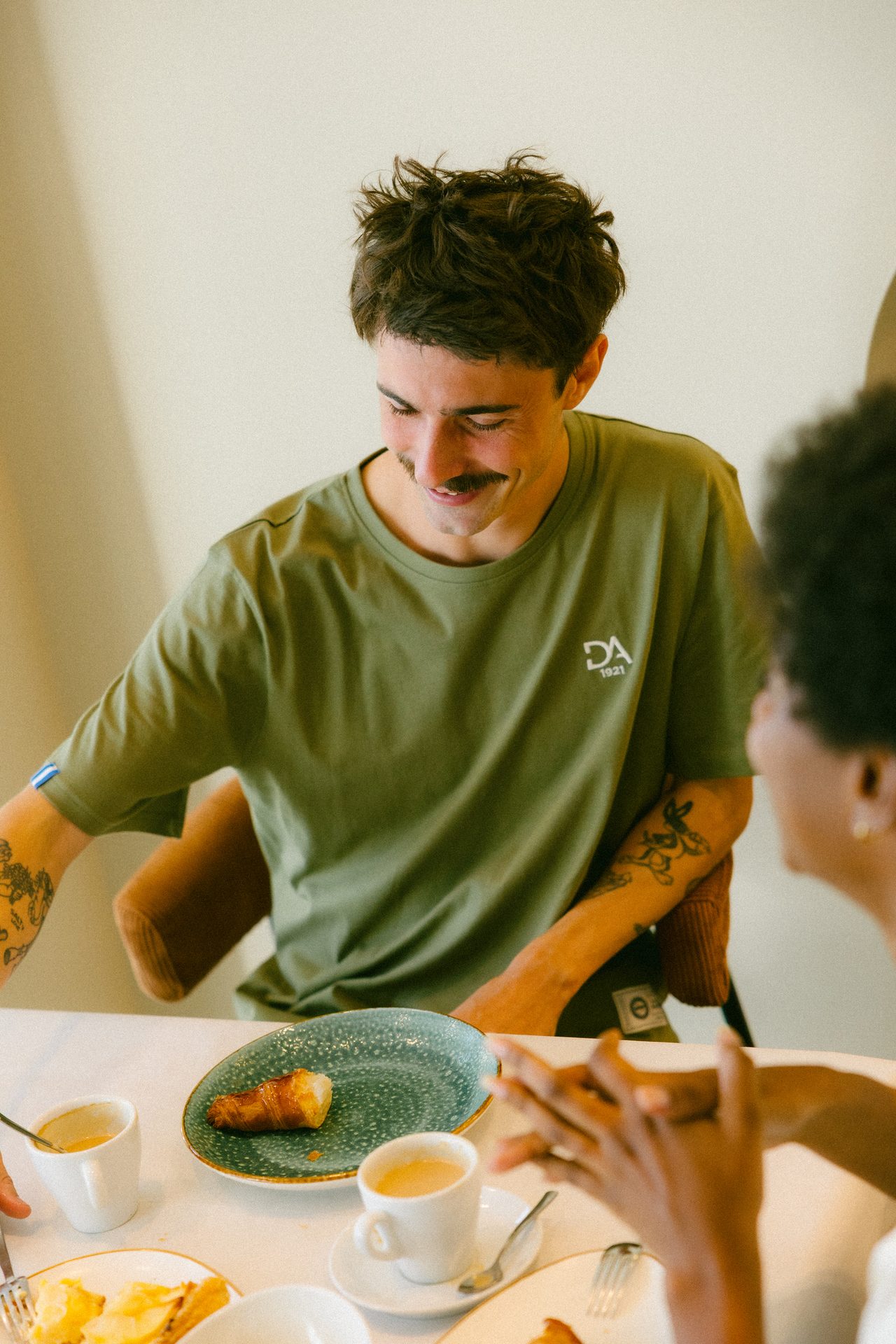 Smiling man with mustache and green shirt at breakfast table with another person.