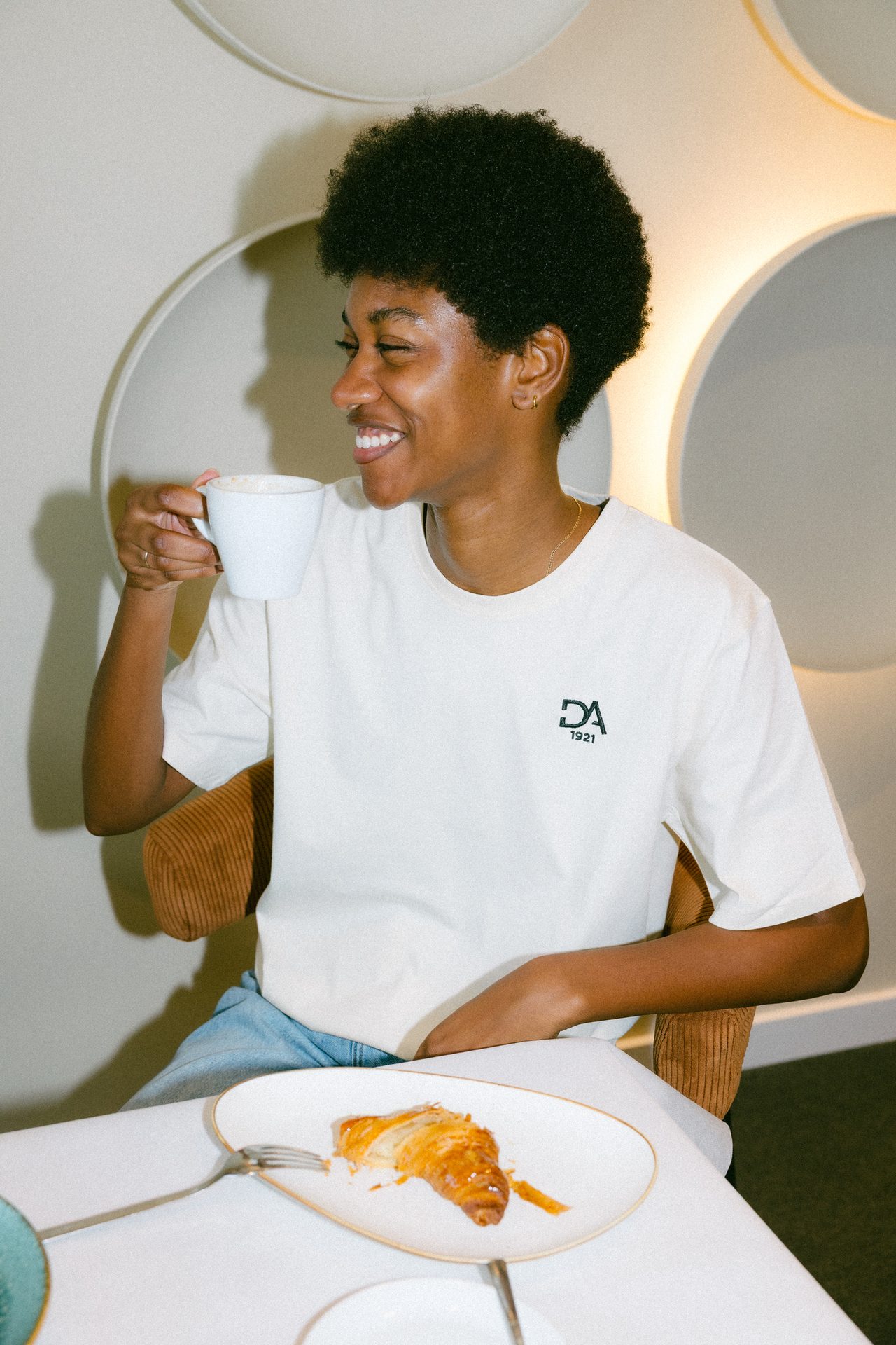 Smiling person sips coffee; pastry on table.