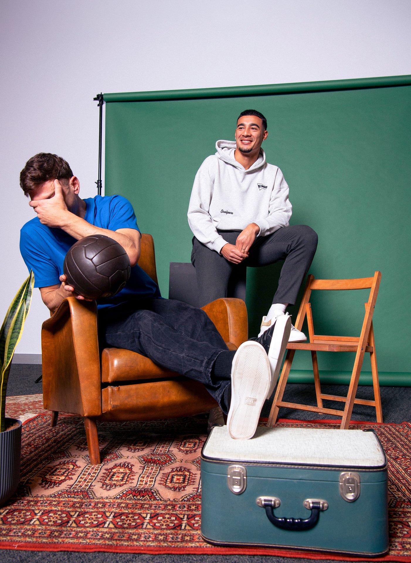 Two men, one looking away, one smiling, pose in a studio with a green backdrop, armchair, and suitcase.