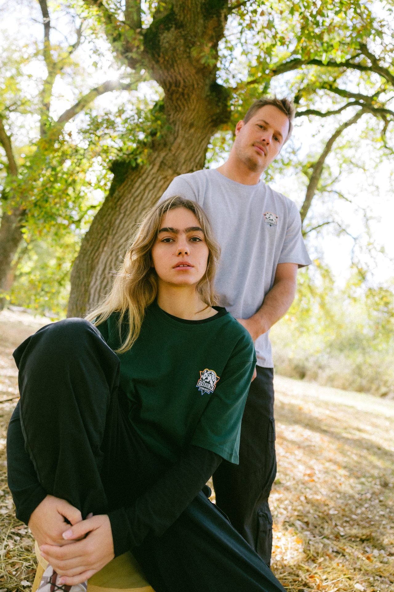 Two people, a woman in green and a man in gray, pose outdoors under trees.
