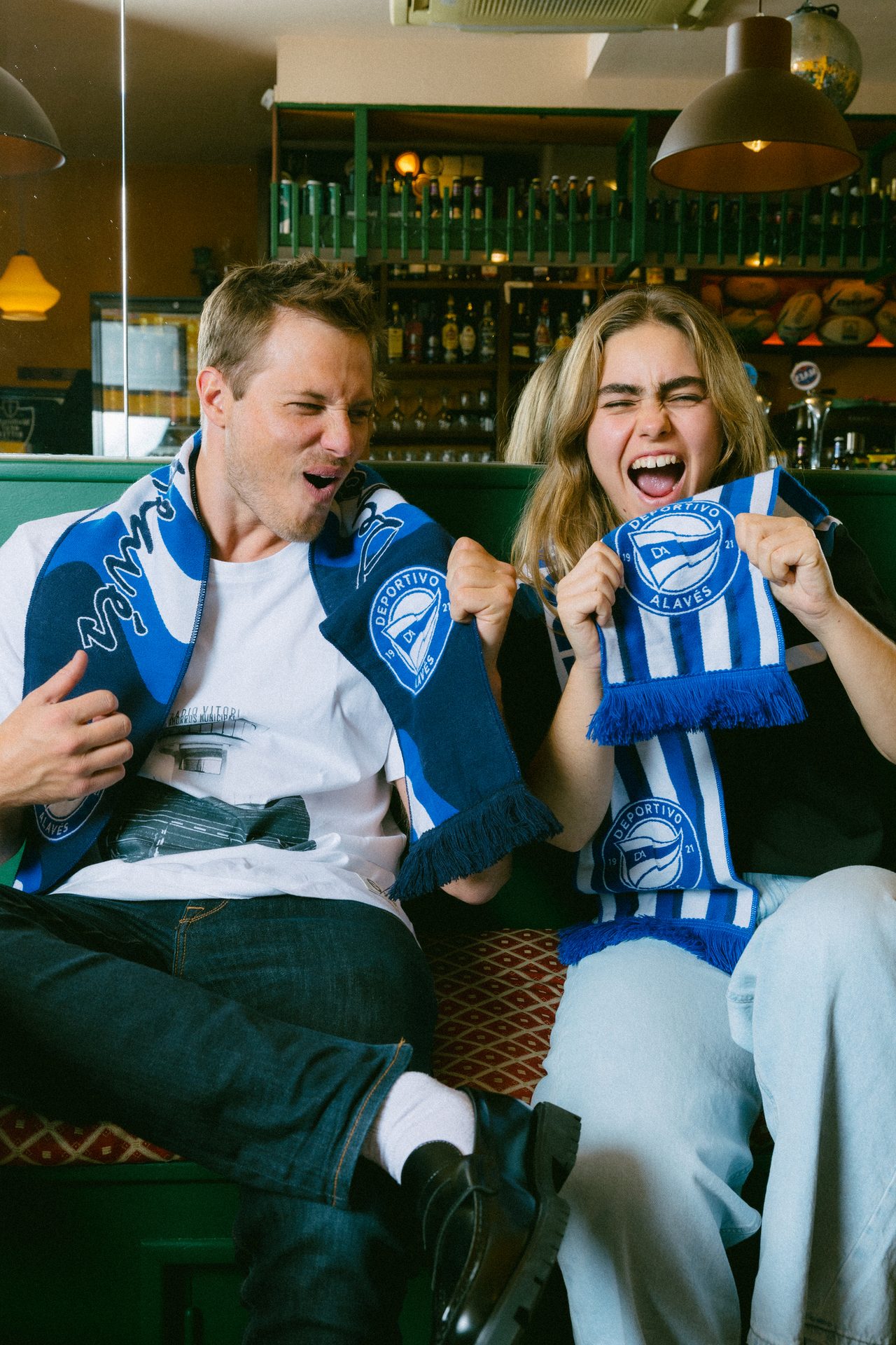 Two excited fans in a bar, wearing Deportivo Alavés scarves.