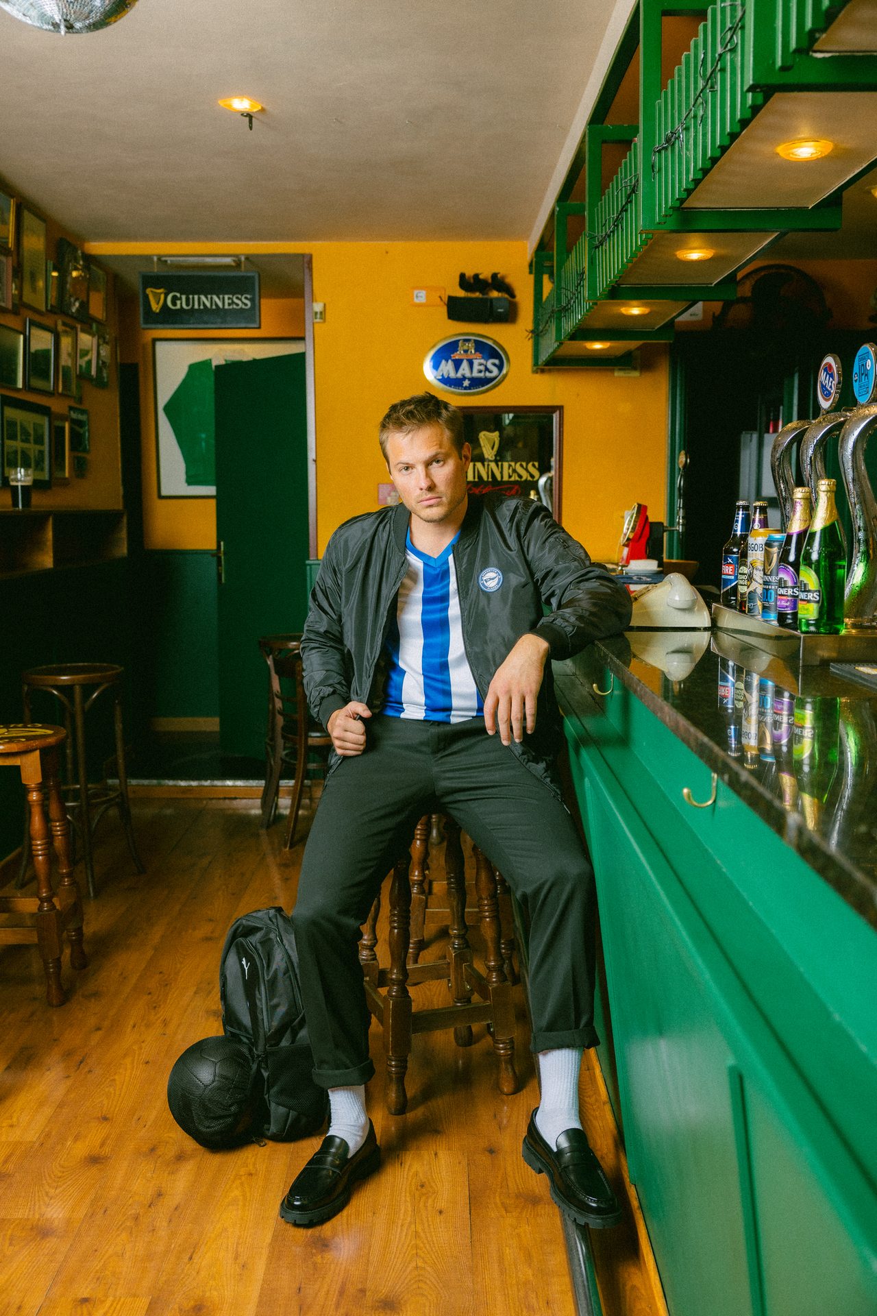 Man in a striped shirt and black jacket sits at a bar counter with a backpack and soccer ball nearby.