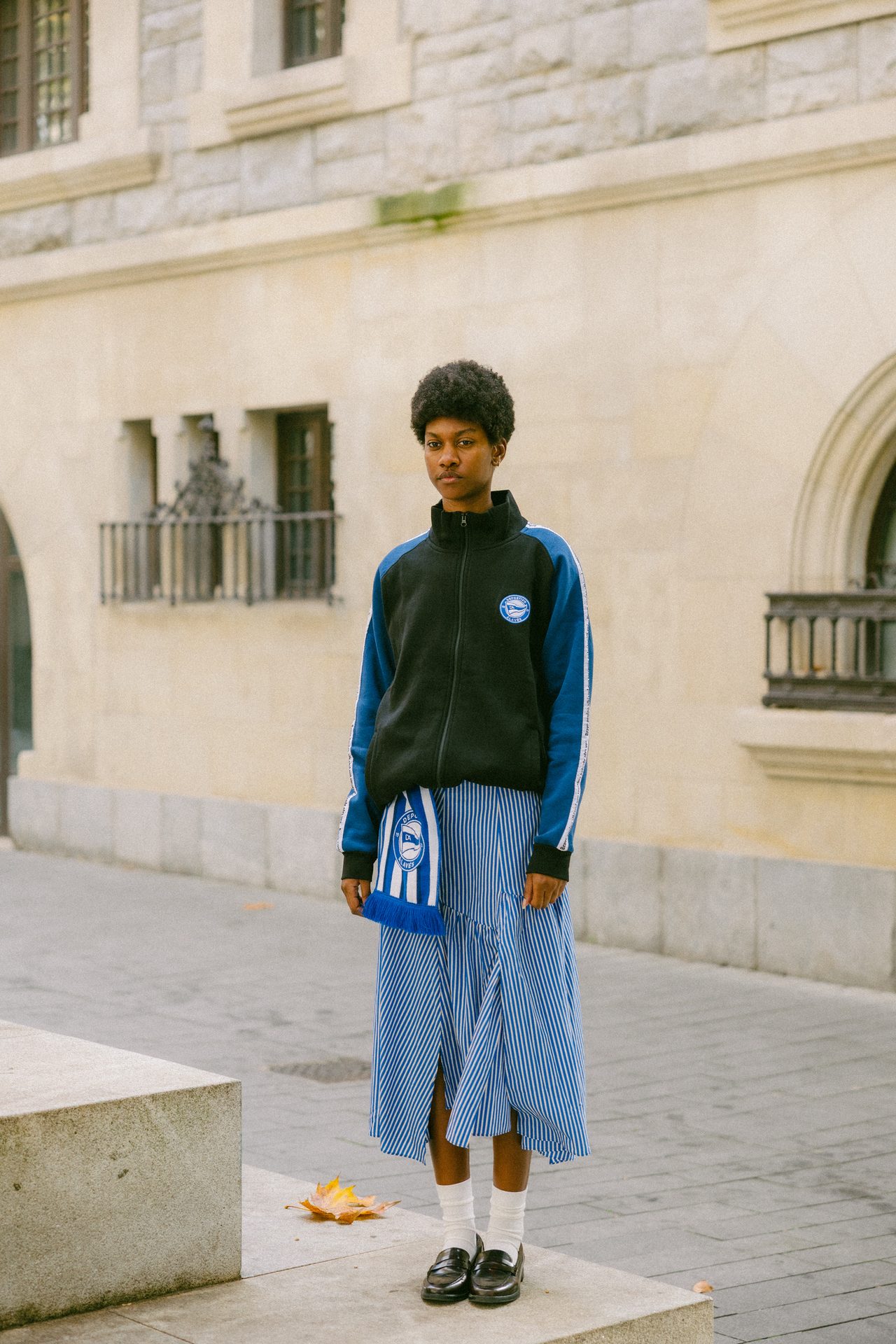 Person in black and blue jacket, striped skirt, white socks, and black loafers, holding a striped scarf.