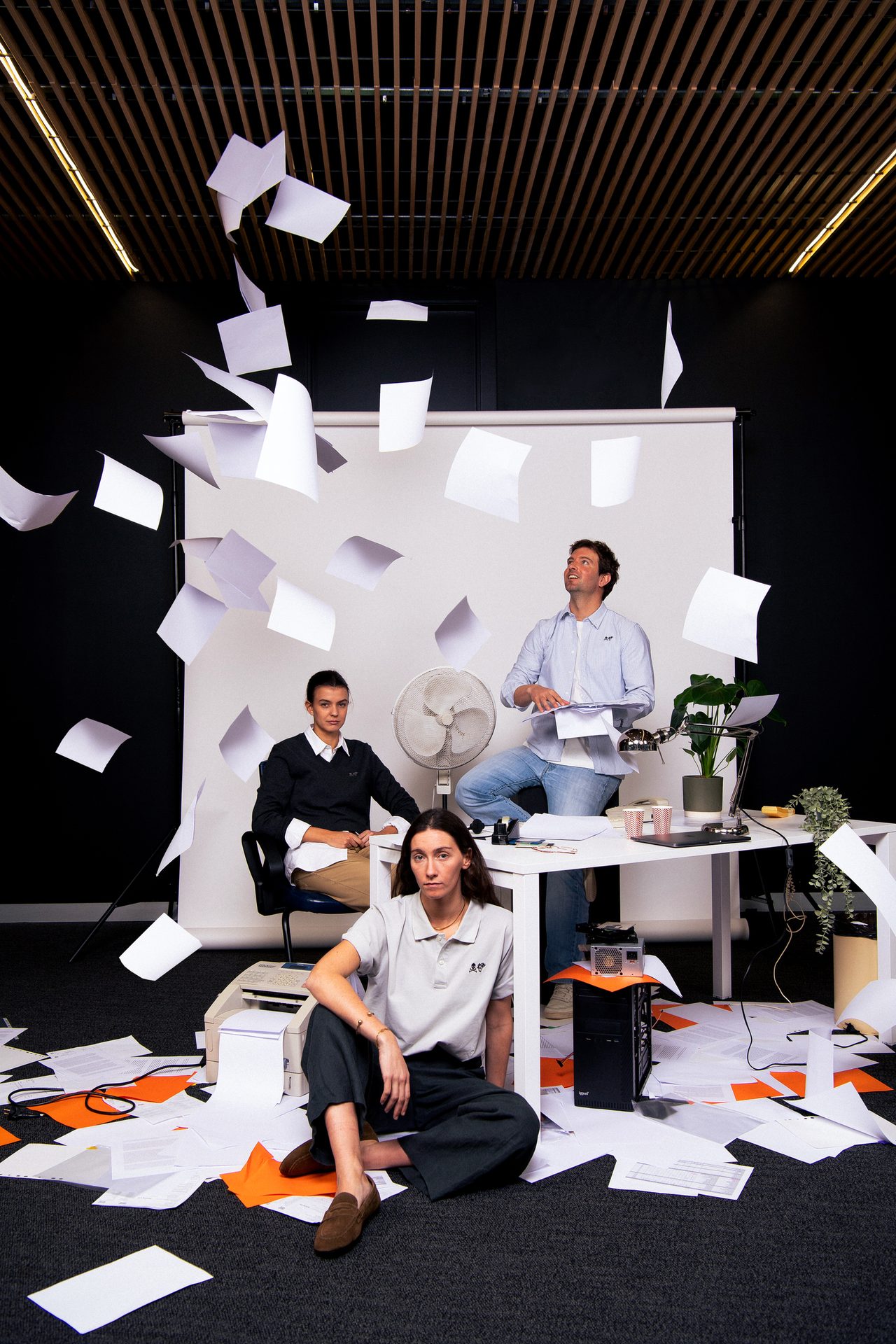 Three colleagues in a chaotic office, with papers flying everywhere and some on the floor.