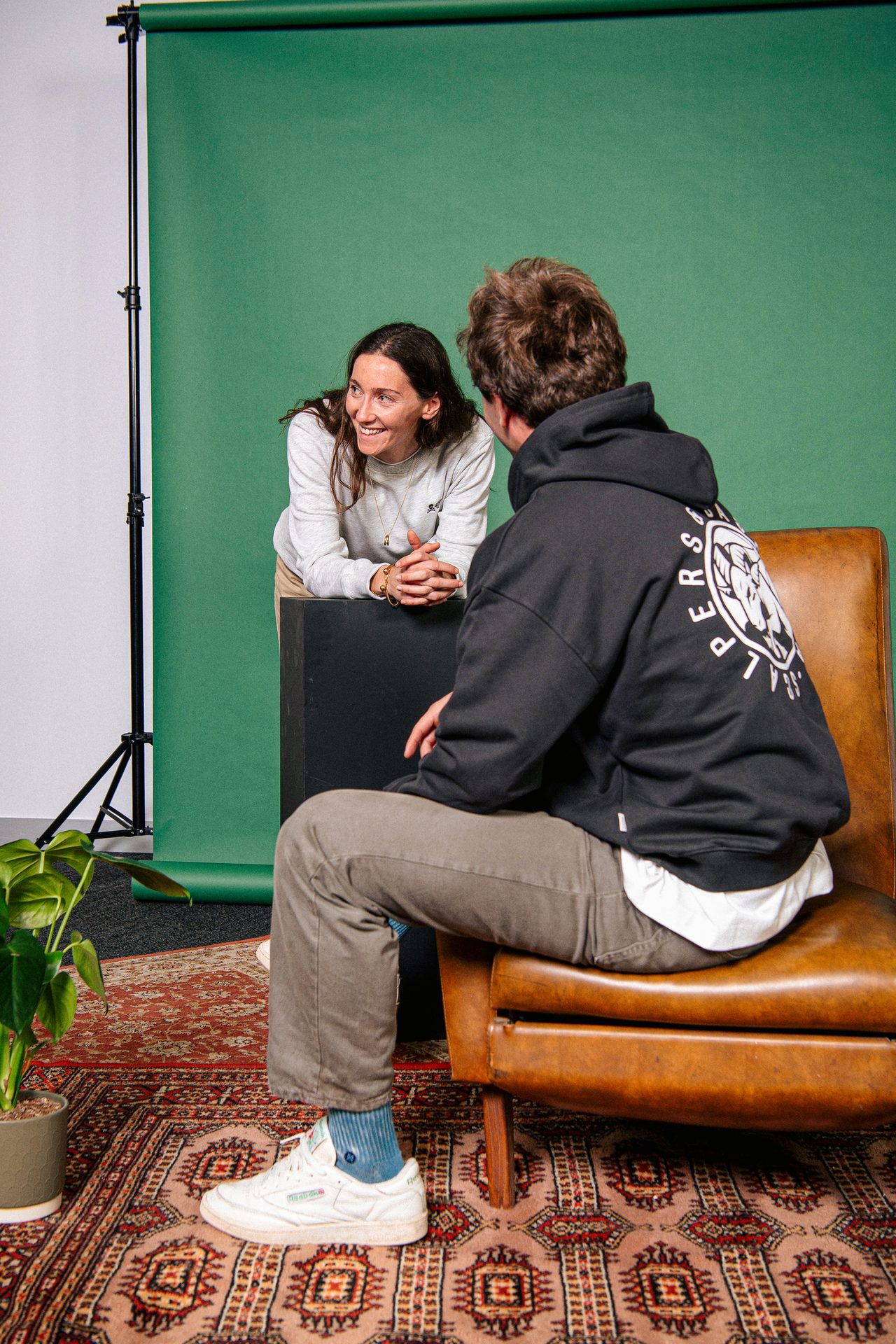 Two people, a woman leaning on a box and a man sitting on a chair, talking in a studio with a green backdrop.