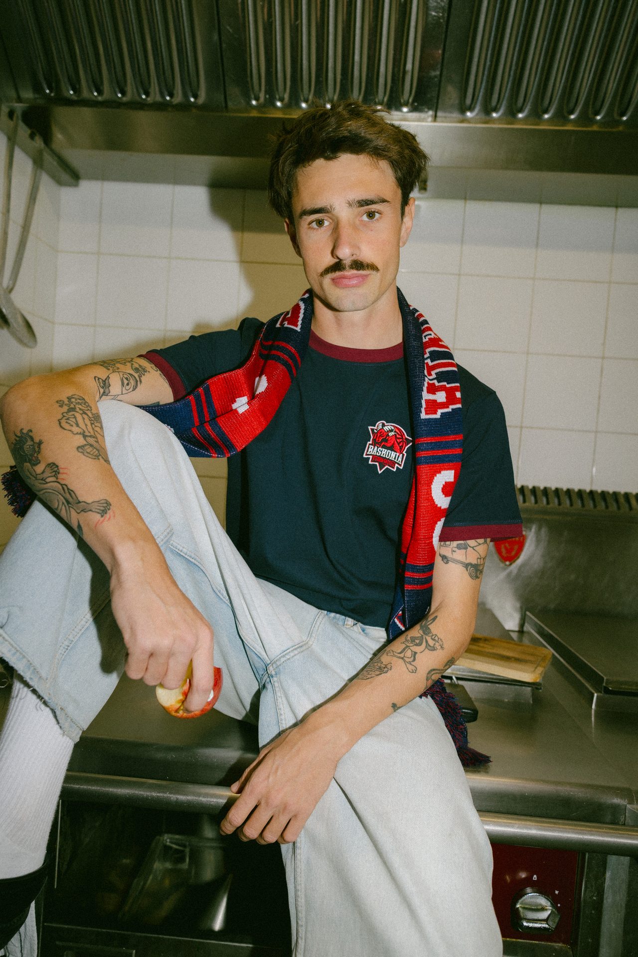 Man with mustache, tattoos, t-shirt, and scarf on a kitchen counter, holding an apple.