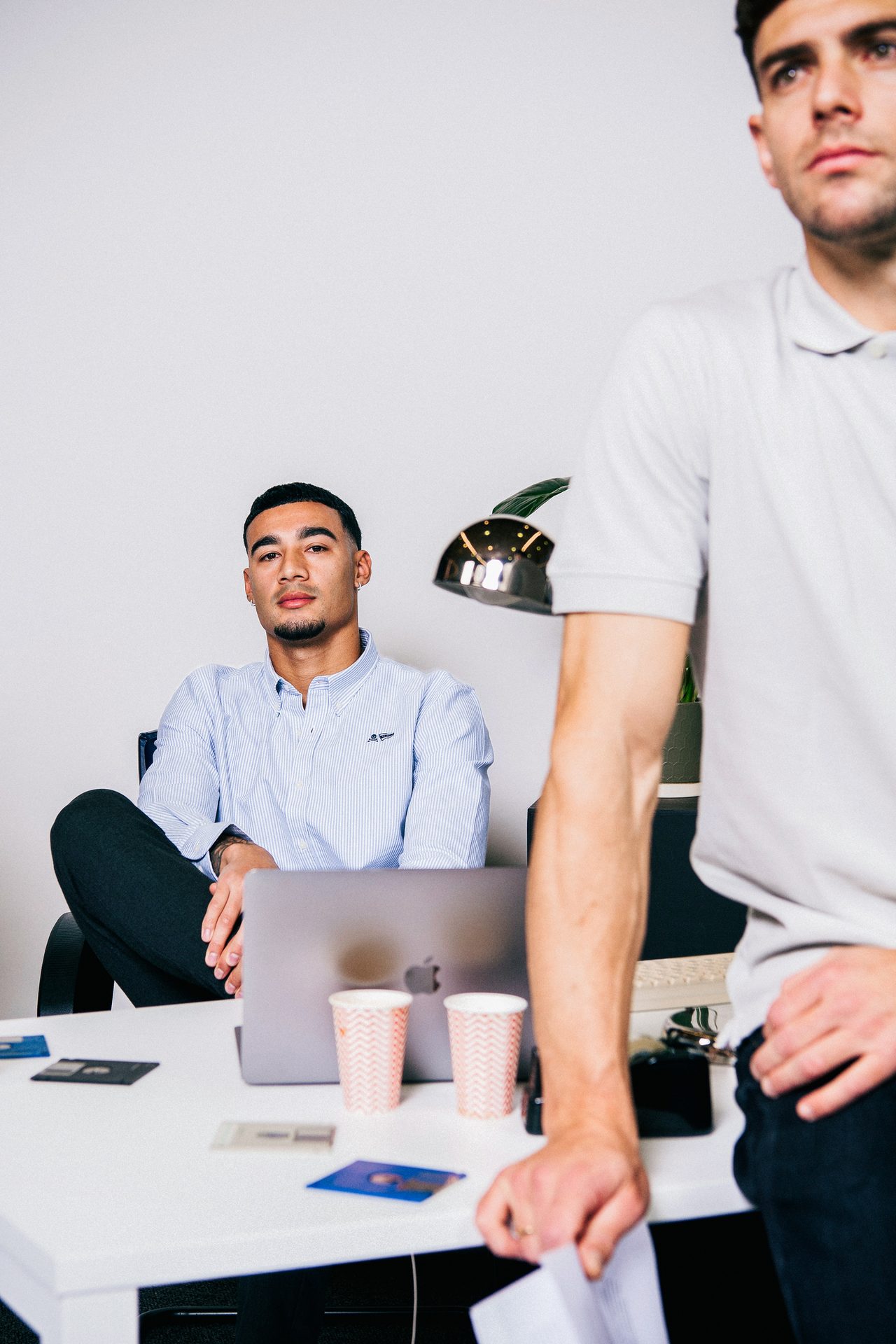 Two young men in an office setting. One sits at a desk with a laptop, another stands beside him.