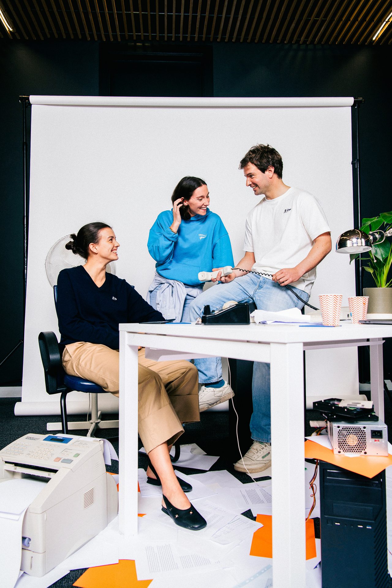Three colleagues laugh and interact playfully in a messy office, with one sitting on a table.