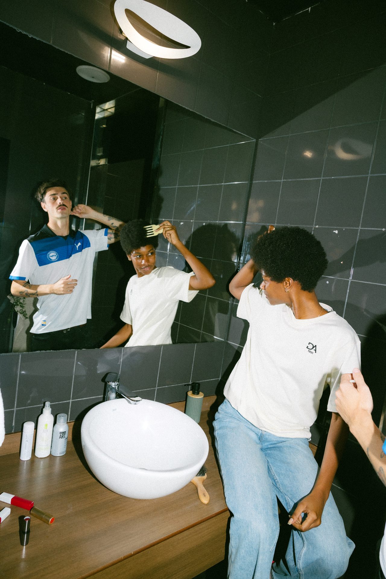 Two people grooming in a bathroom: a man reflected adjusts his hair, a woman combs her afro.