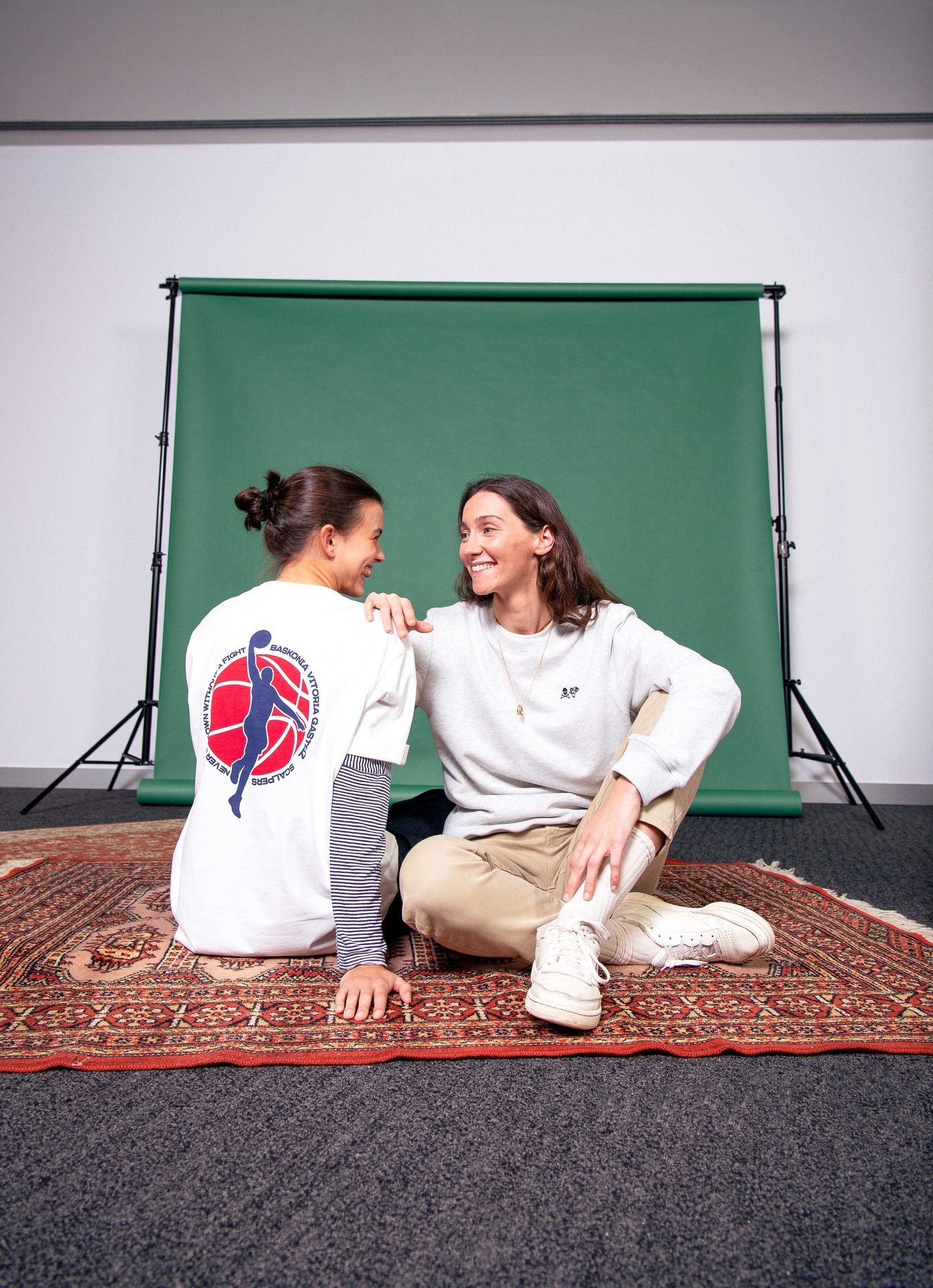 Two women sitting on a rug, facing each other and smiling in front of a green backdrop.