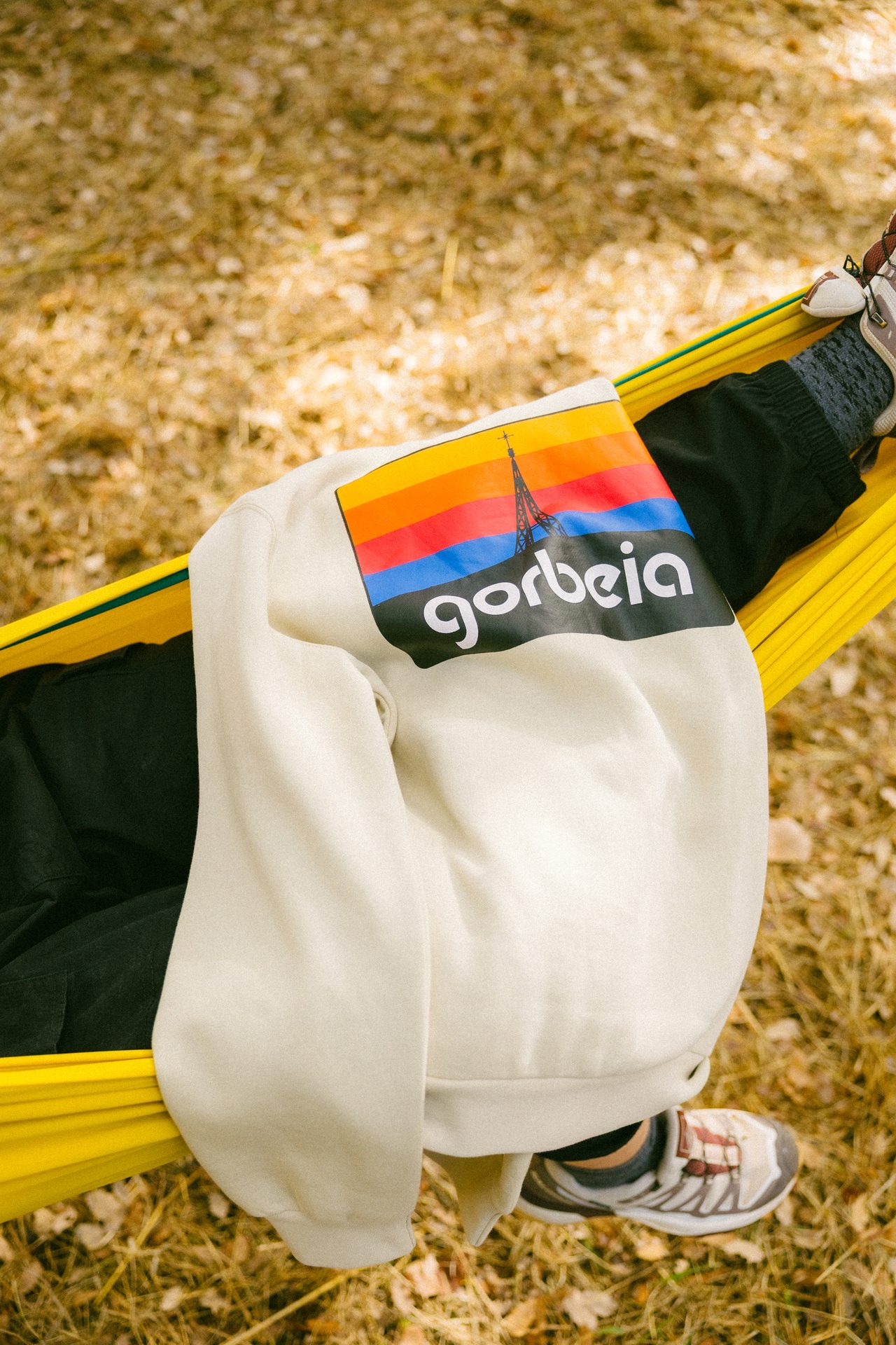 Person relaxing in a yellow hammock, wearing a cream "gorbeia" sweatshirt, black pants, and hiking shoes on autumn leaves.