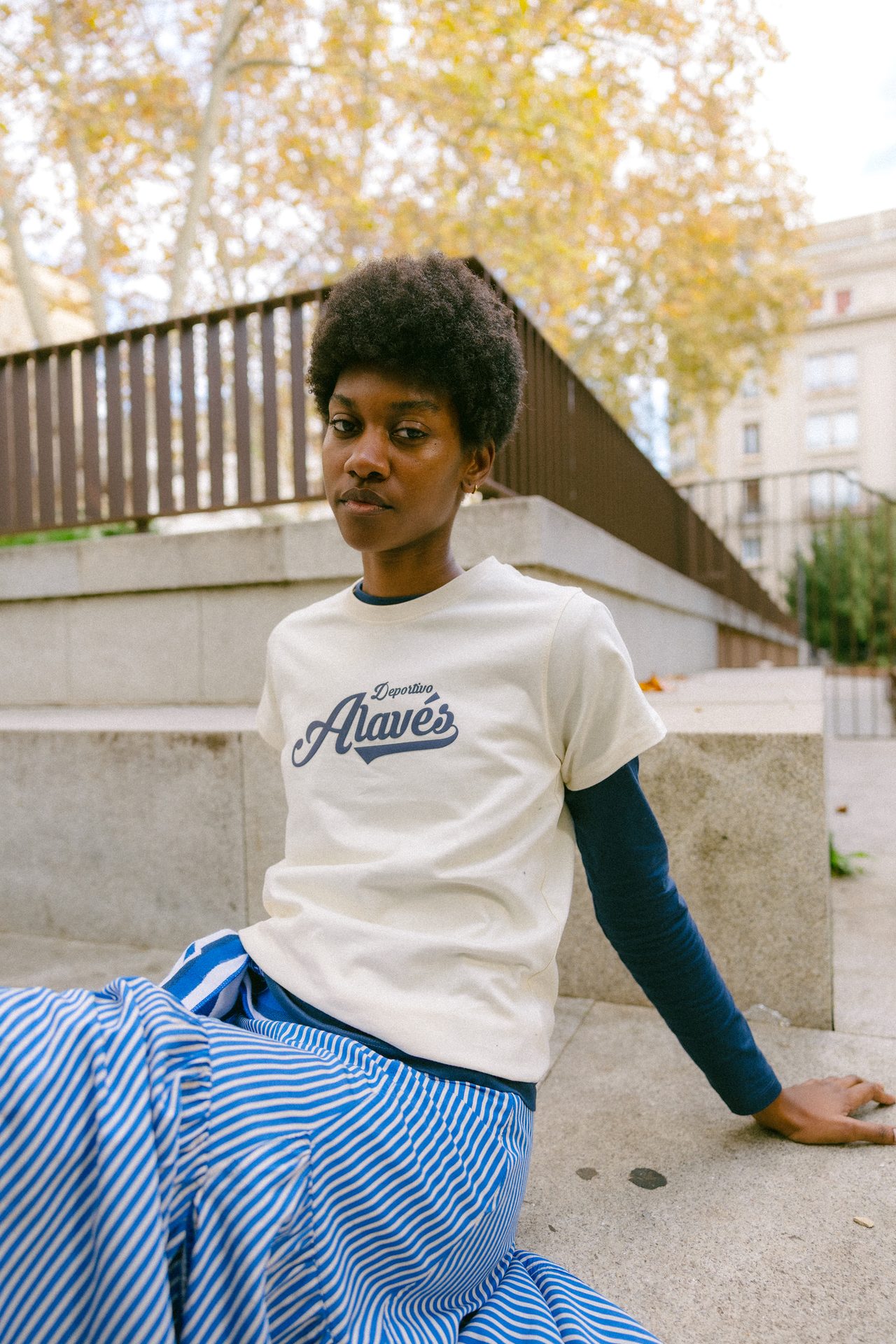 A person with afro hair, wearing a cream "Deportivo Alavés" tee and striped pants, sits outdoors.