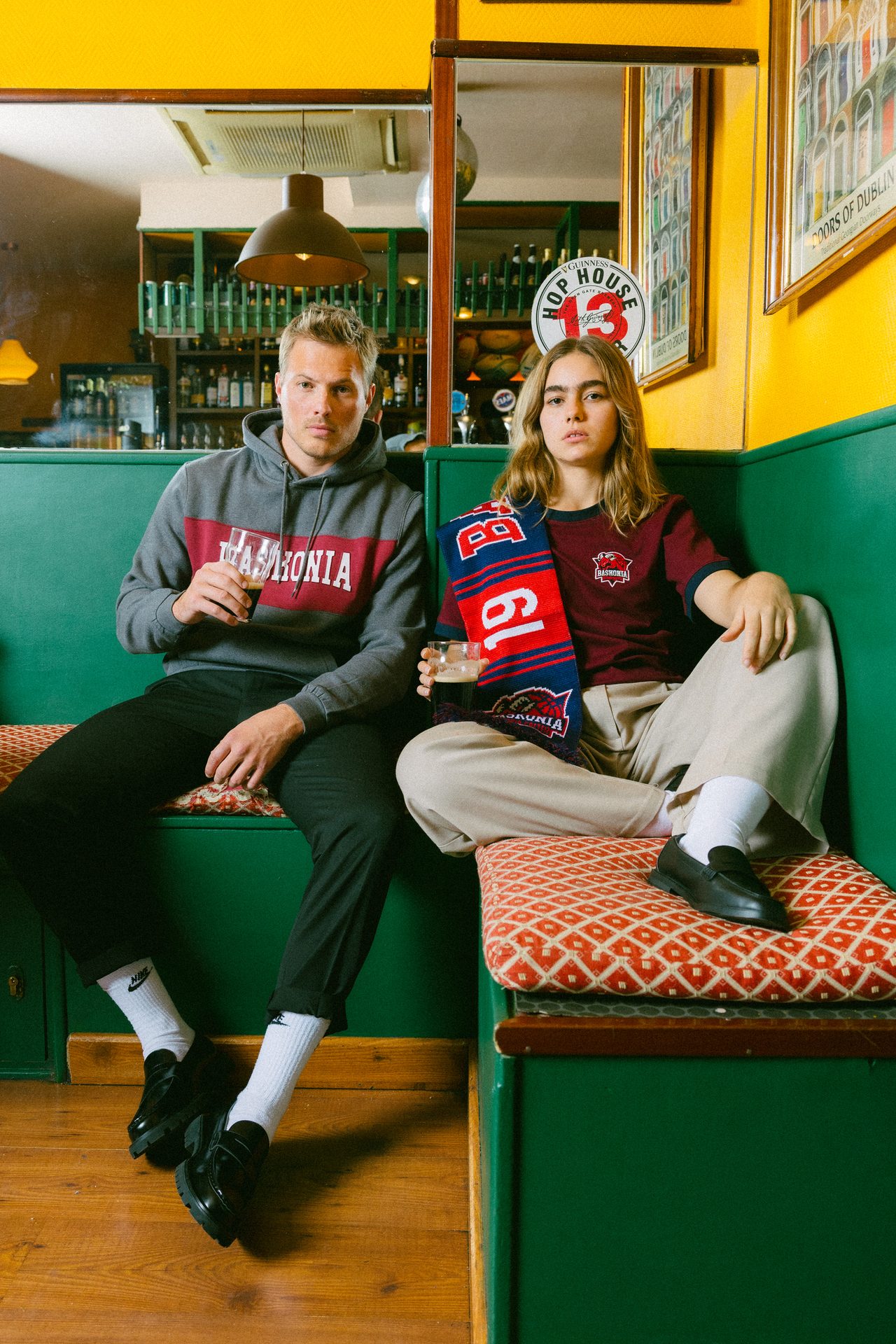 Man and woman sit in a pub booth with drinks. He wears a grey hoodie, she has a burgundy top and scarf.