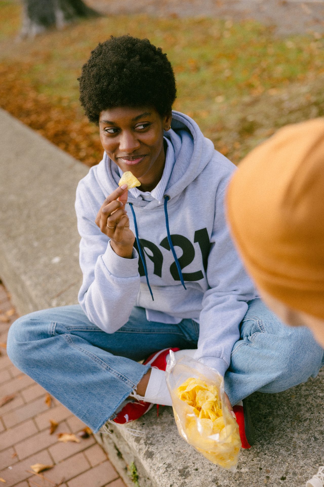 A person in a grey hoodie sits outdoors, eating chips and looking at another person.