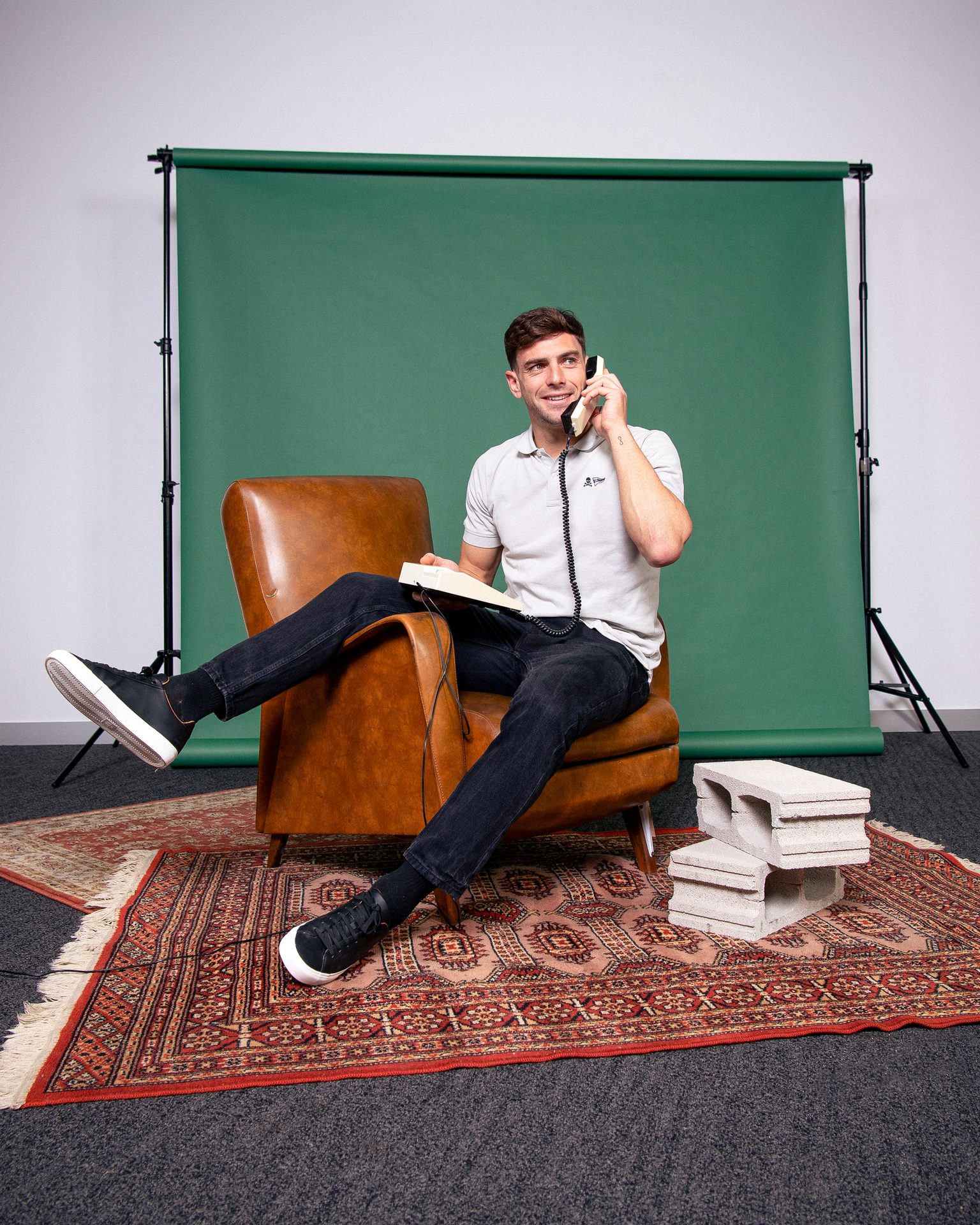 A man in an armchair talking on a retro telephone in front of a green backdrop.