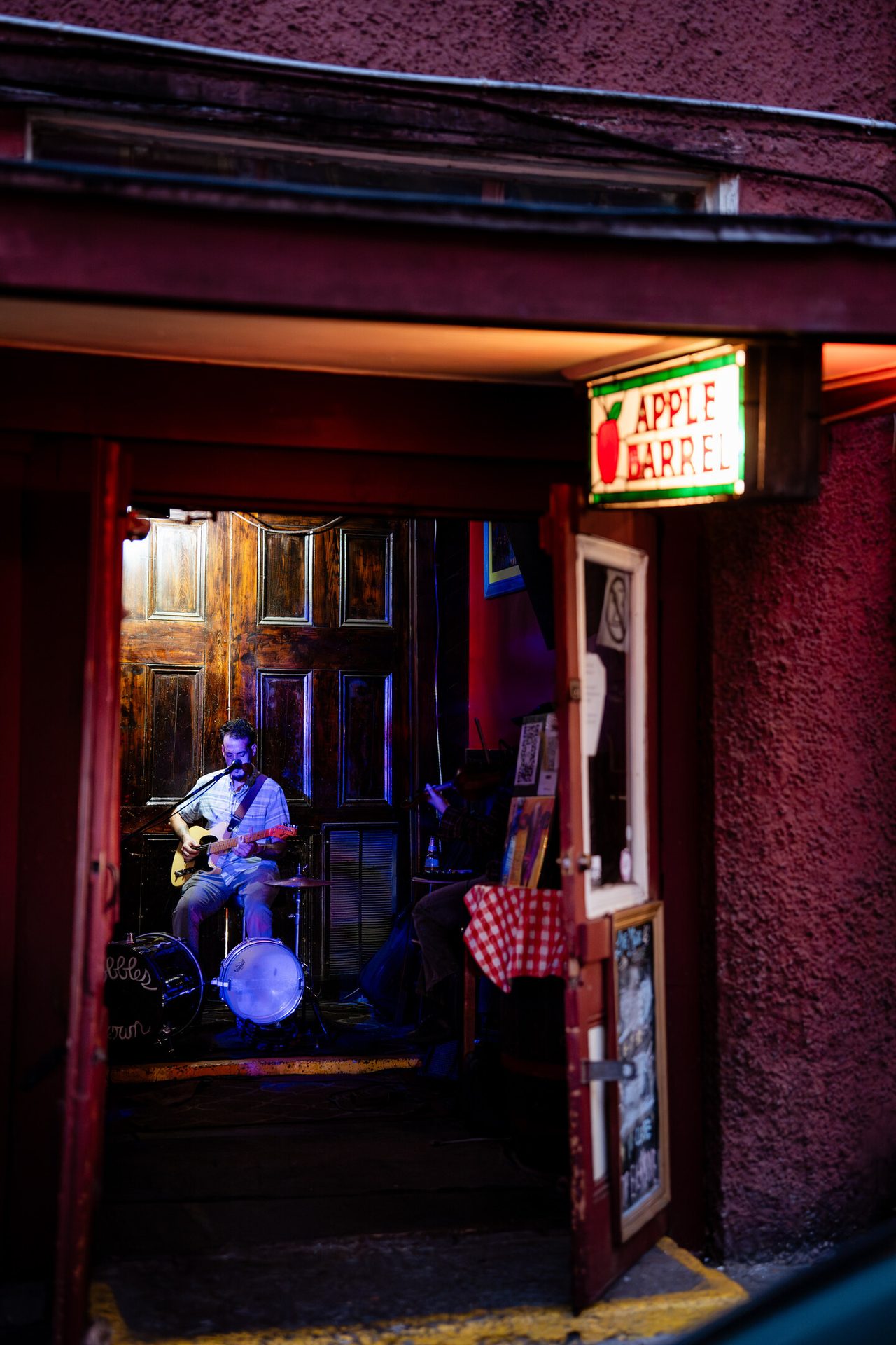 Musician playing guitar in a dimly lit doorway under an "Apple Barrel" sign.