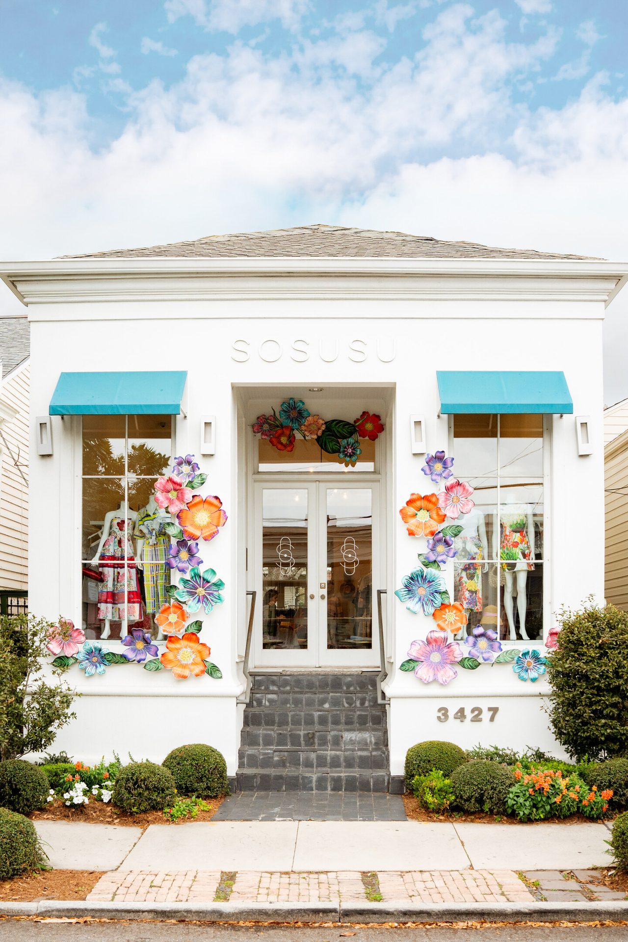 White shopfront decorated with colorful artificial flowers around windows and door, with "SOSUSU" sign.