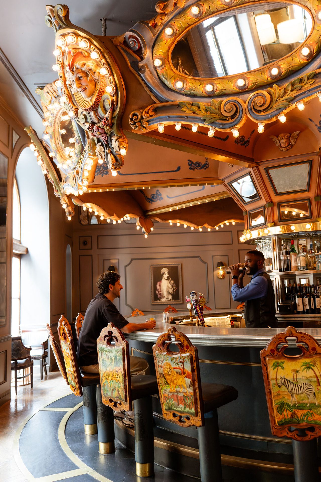 A man sits at a carousel-themed bar with intricate details, while a bartender mixes a drink.