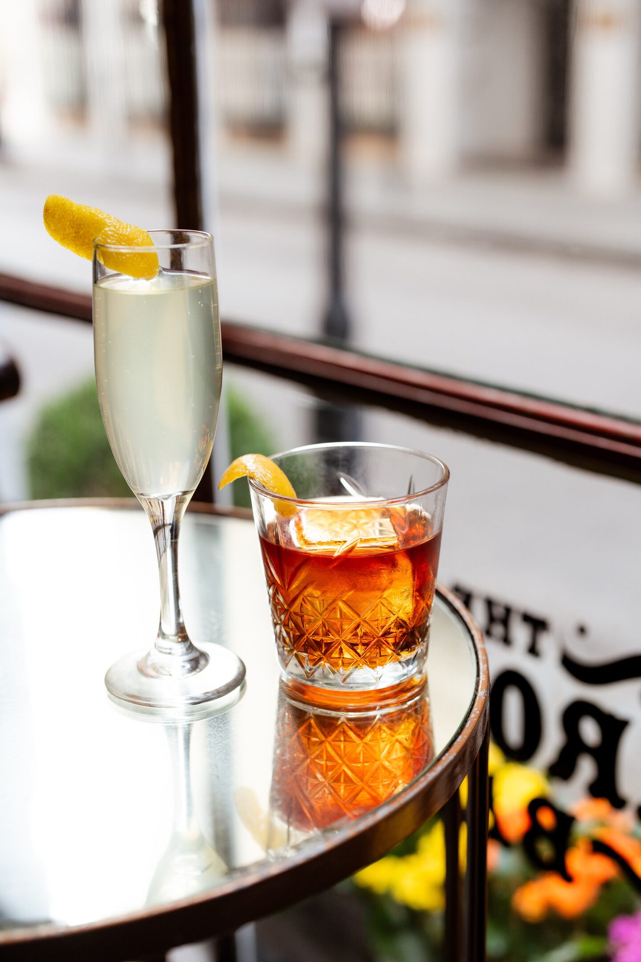 Two cocktails, one light in a flute and one dark in a rocks glass, with citrus garnishes on a reflective table.