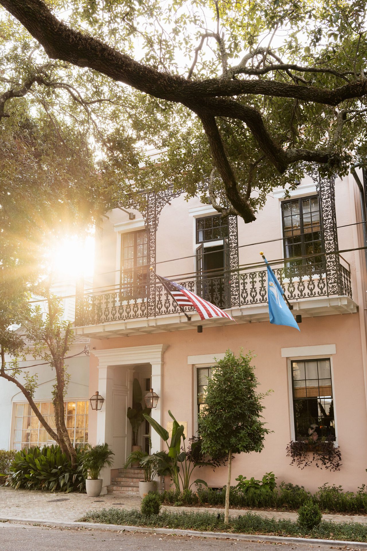 A charming light pink building with an ornate balcony, flags, lush greenery, and dappled sunlight.