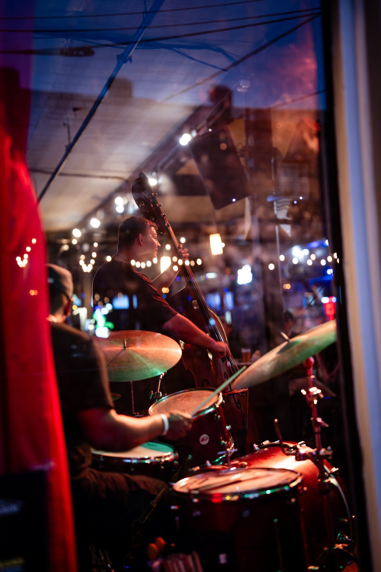 Two musicians, a bassist and a drummer, perform live music seen through a window with city reflections.