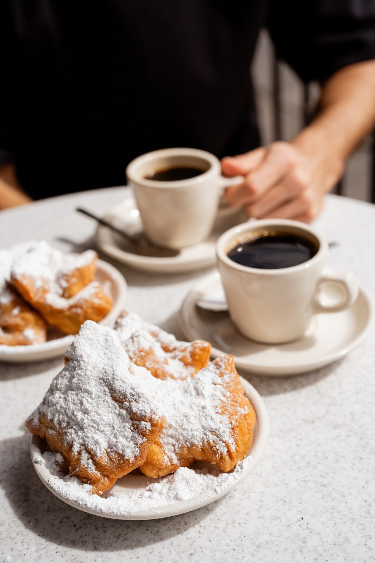 A table with several plates of powdered sugar beignets and two cups of black coffee, with a hand holding one.