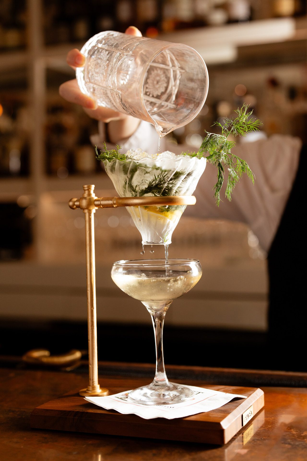 A bartender pouring liquid through a botanical-filled filter into a coupe glass on a brass stand.