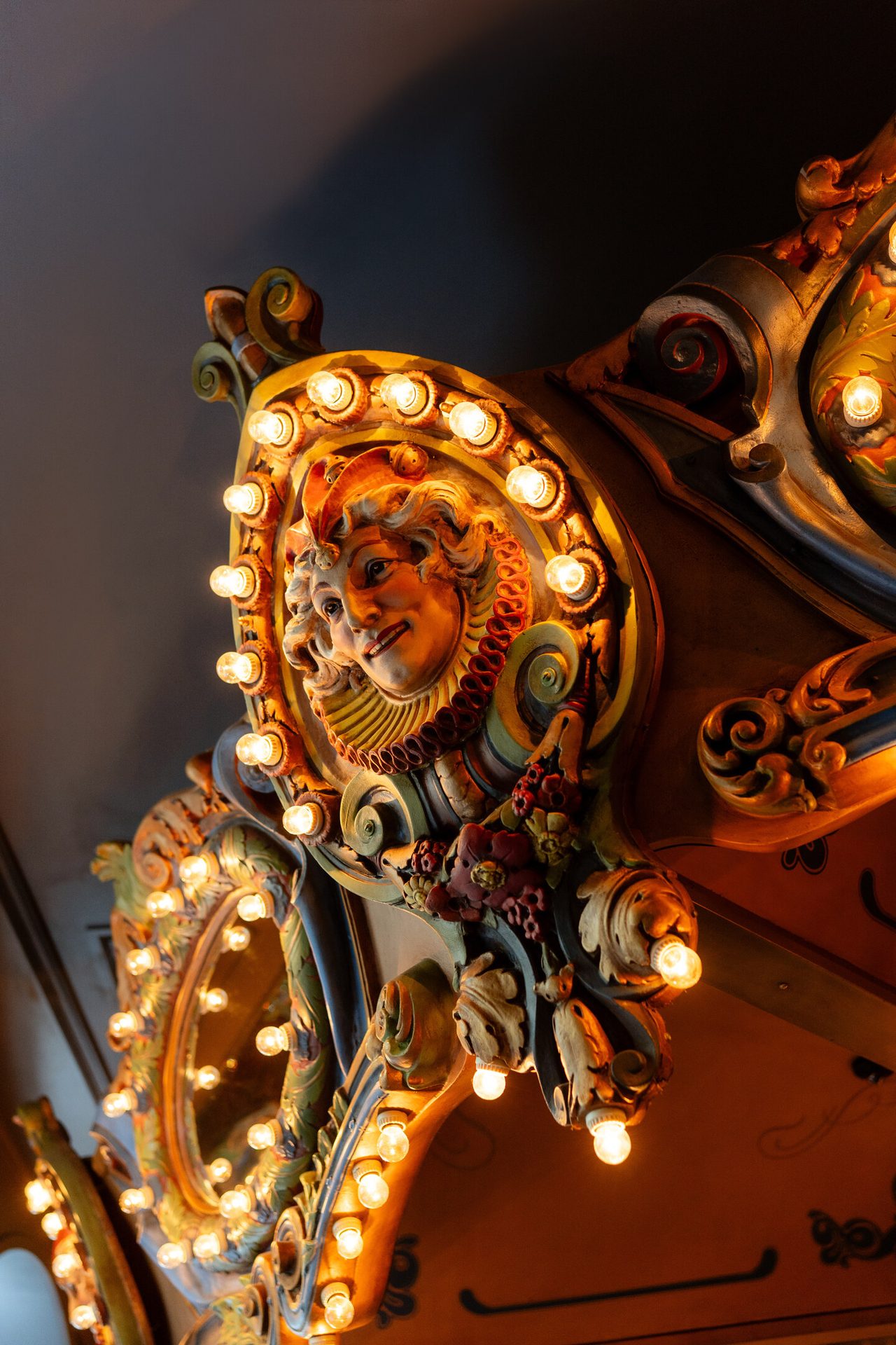 Ornate carved face with a theatrical expression, surrounded by glowing lights on a vintage carousel.