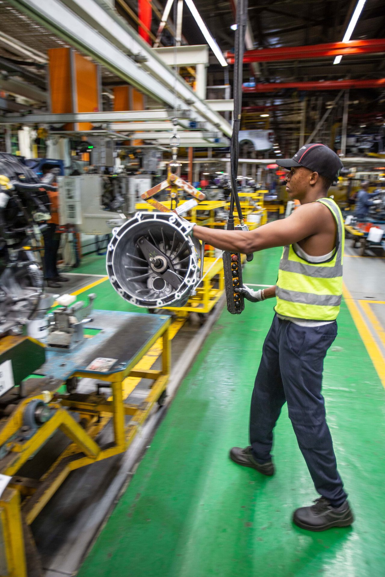 A worker on an assembly line uses a hoist to move a large engine part; motion blur shows activity.