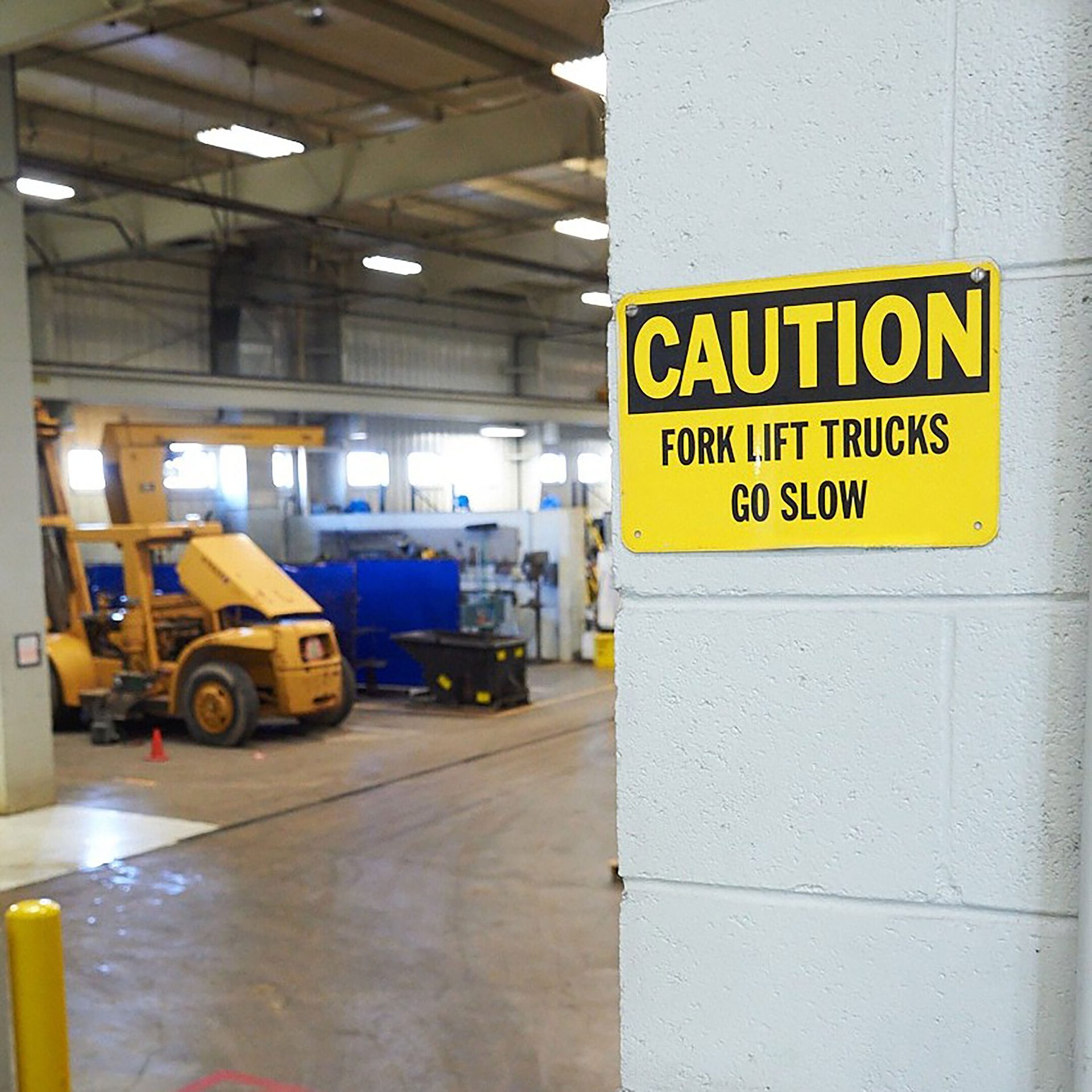 Yellow caution sign "FORK LIFT TRUCKS GO SLOW" on a white wall, with a forklift in a warehouse.