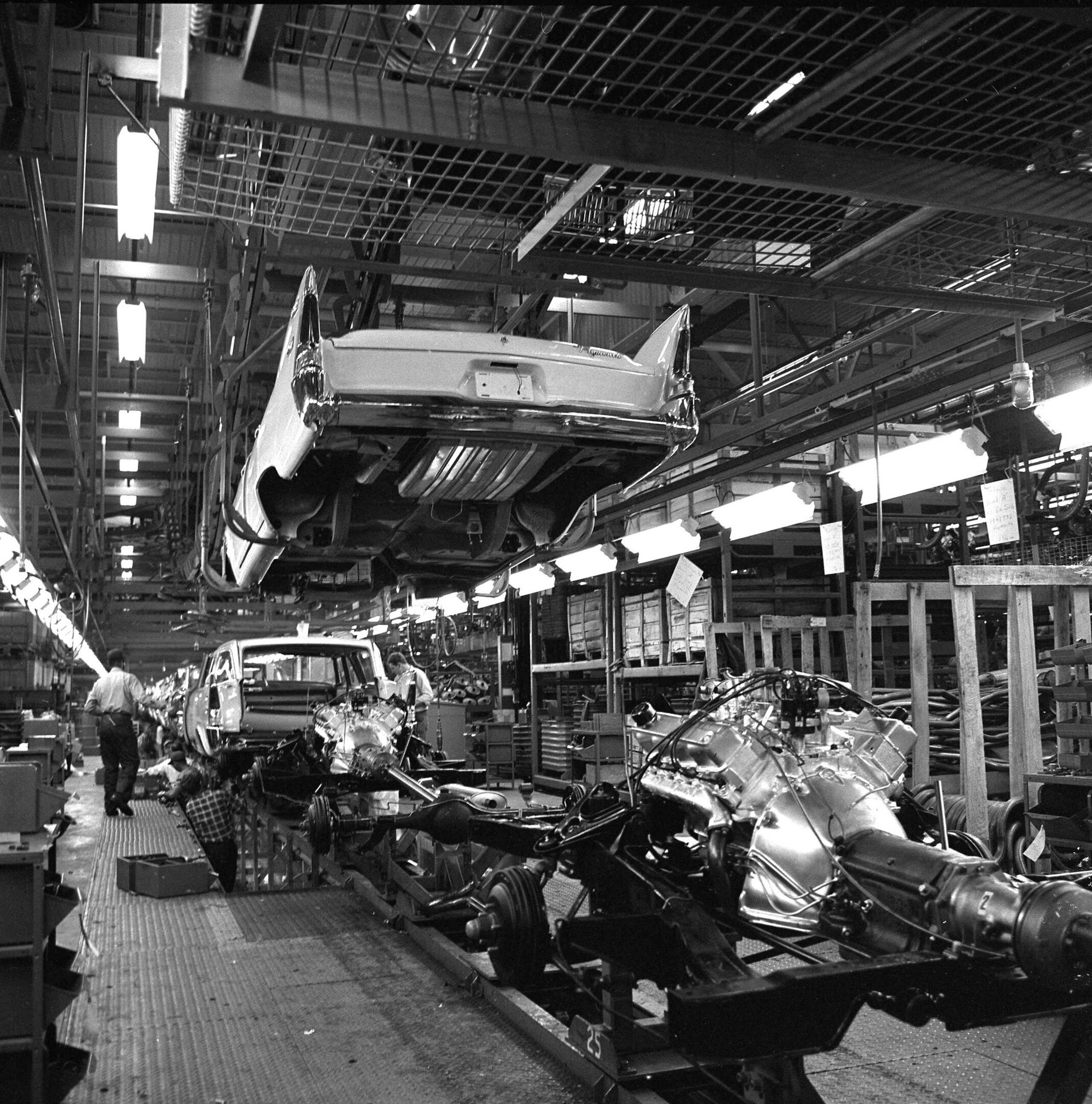 Black and white image of a busy car assembly line with car bodies on an overhead conveyor and chassis below.