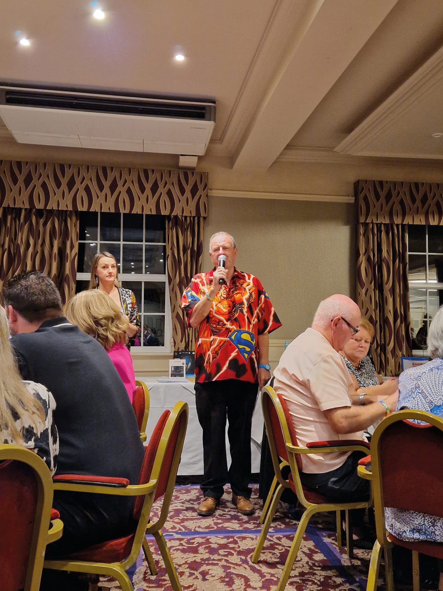 Man in a Superman shirt speaks into a microphone, with a woman standing behind him, in a room with seated guests.