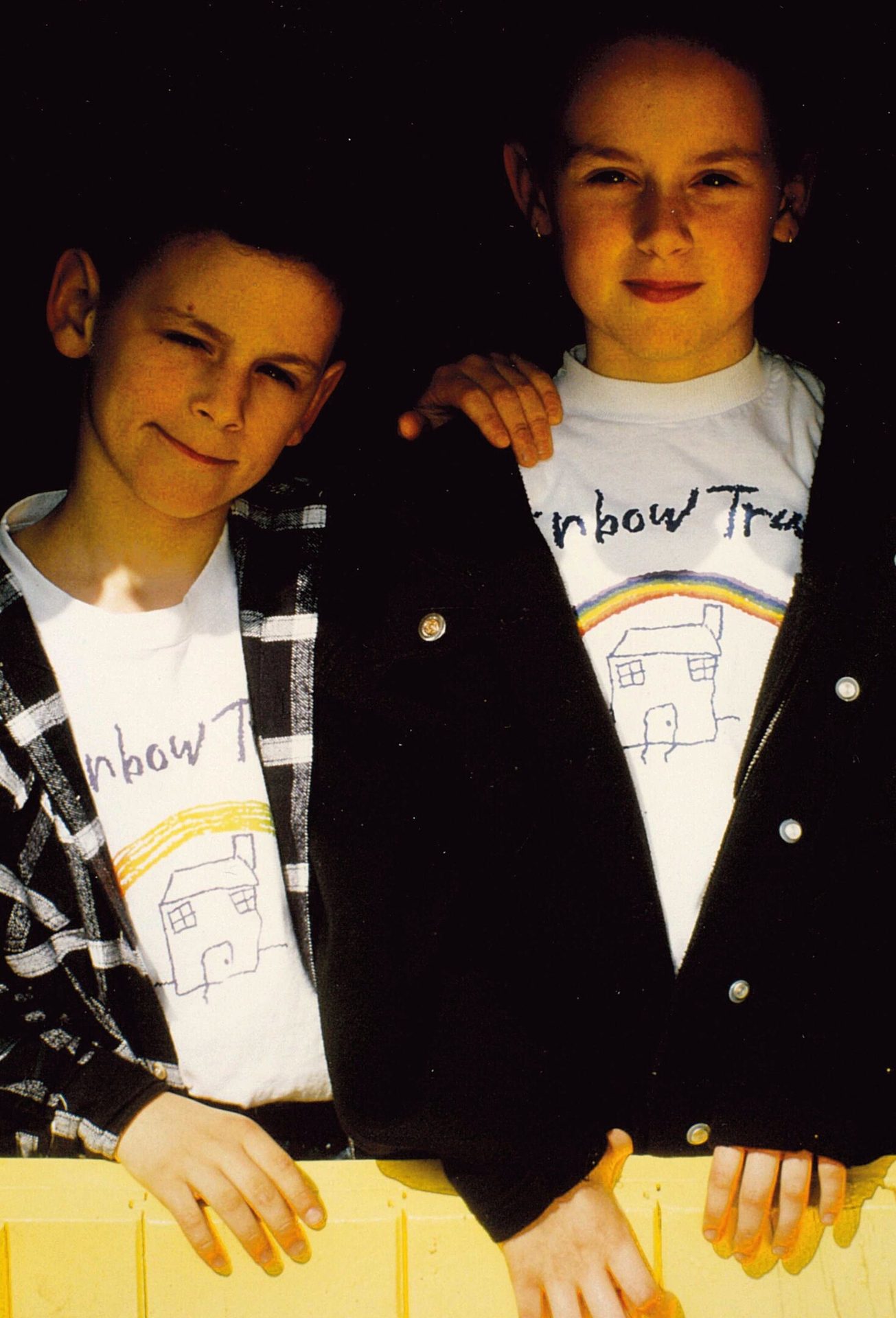 Two children in 'Rainbow Trust' t-shirts with a house and rainbow design, smiling.