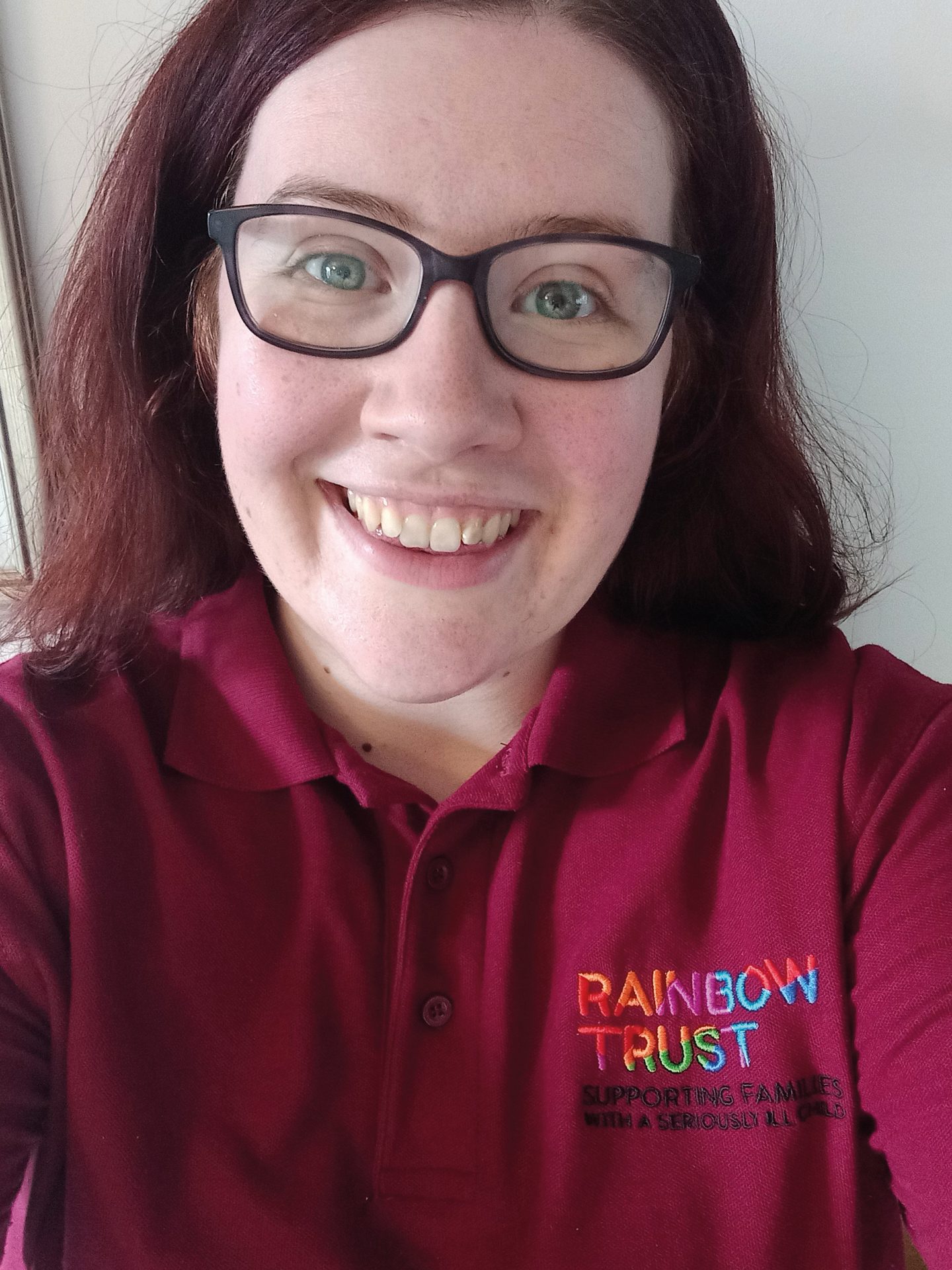 Smiling woman in burgundy Rainbow Trust polo shirt with logo: 'Supporting families with a seriously ill child'.