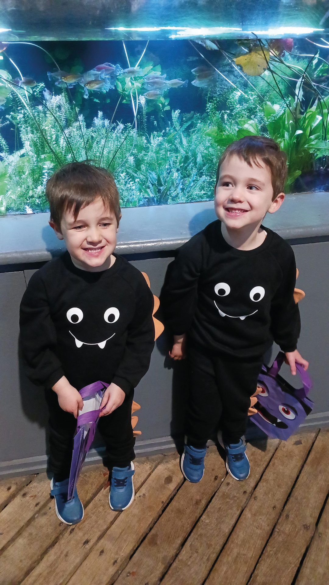 Two smiling boys in black monster outfits stand in front of an aquarium, holding trick-or-treat bags.