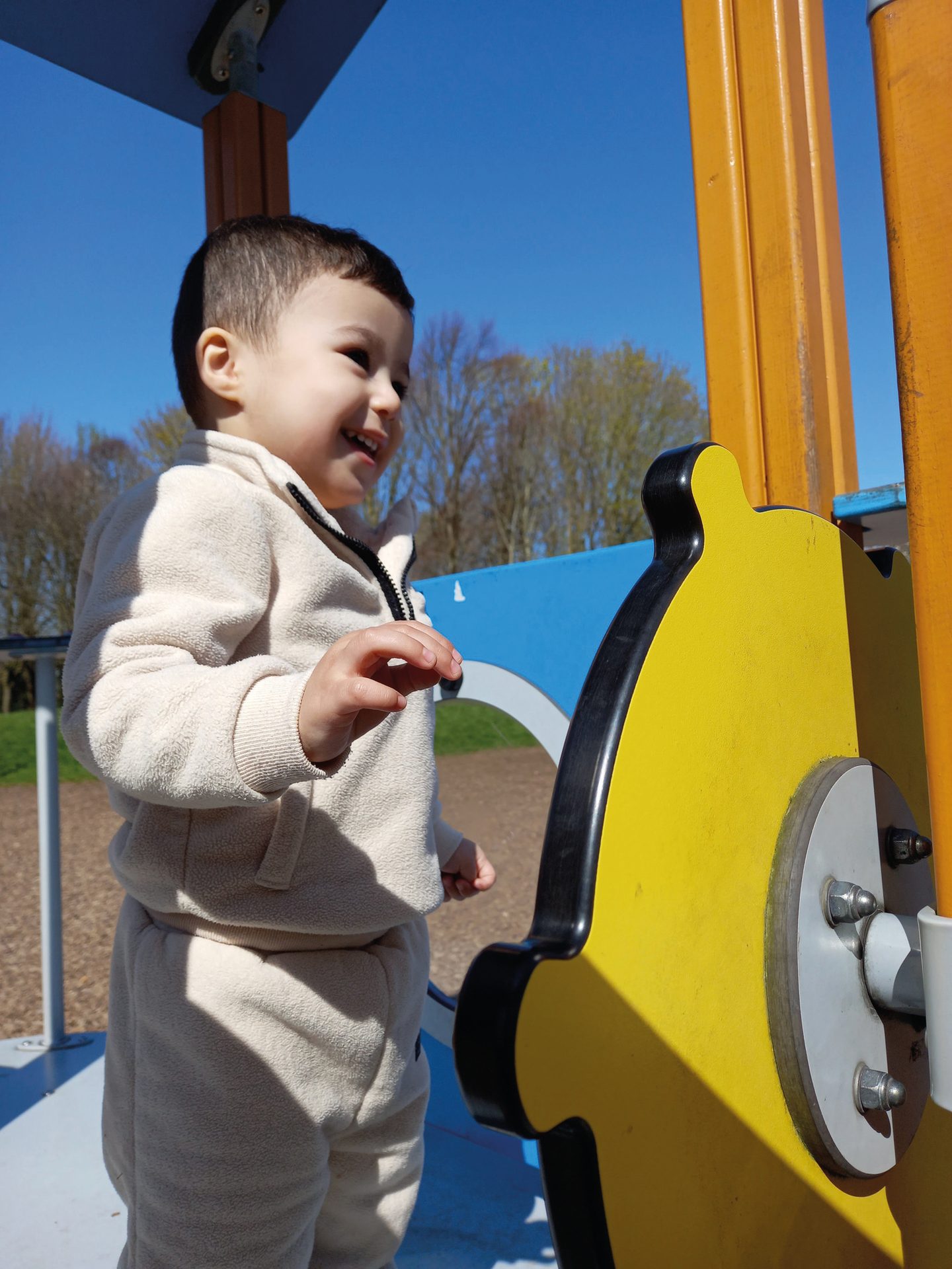 Smiling child in cream tracksuit plays on a yellow playground structure under a blue sky.