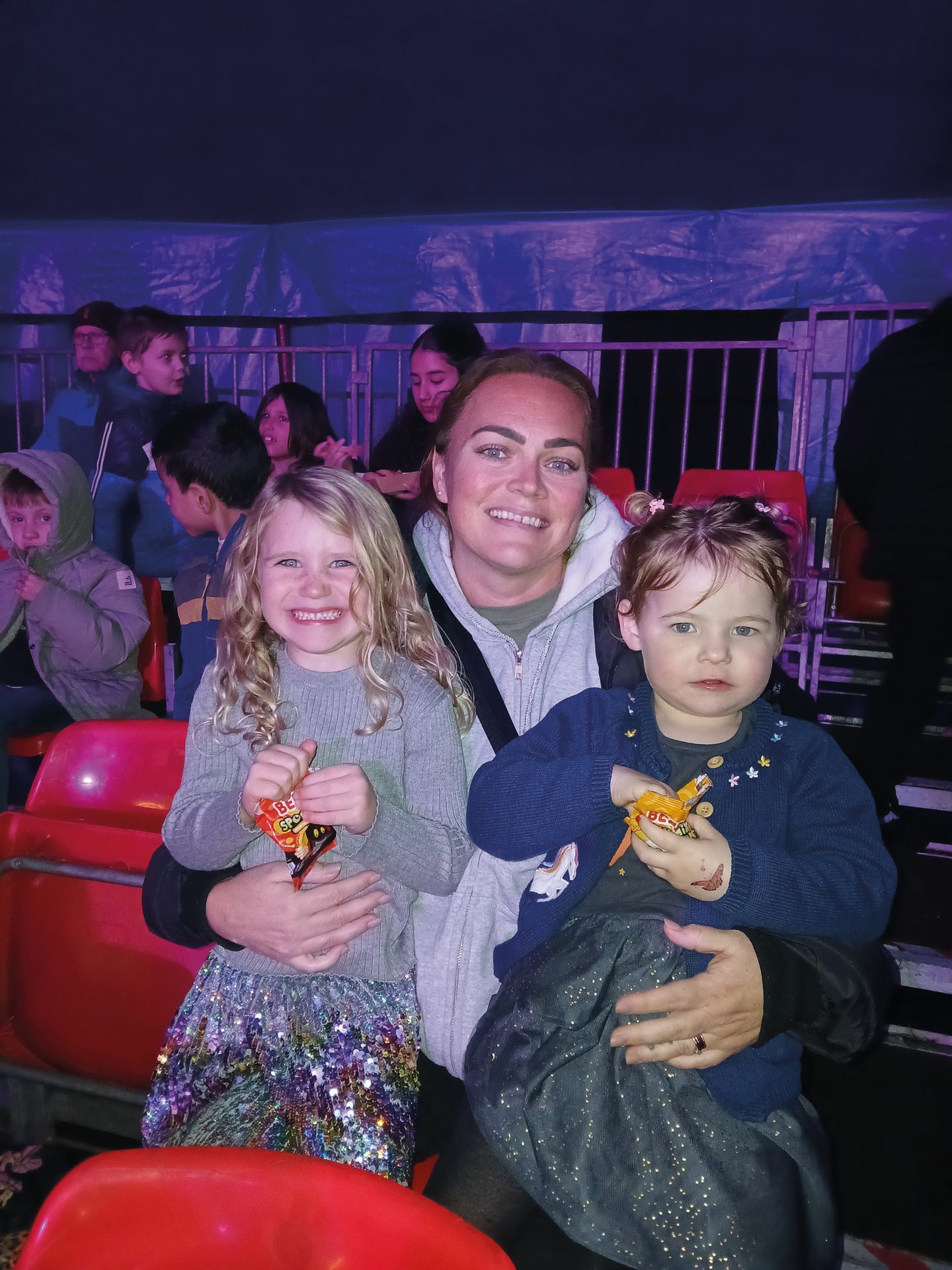 A smiling woman with two young girls, holding candy, seated at an event.