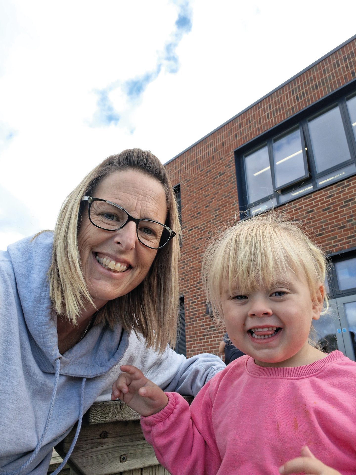 A woman in glasses and a blonde child smile for a selfie outside a brick building.