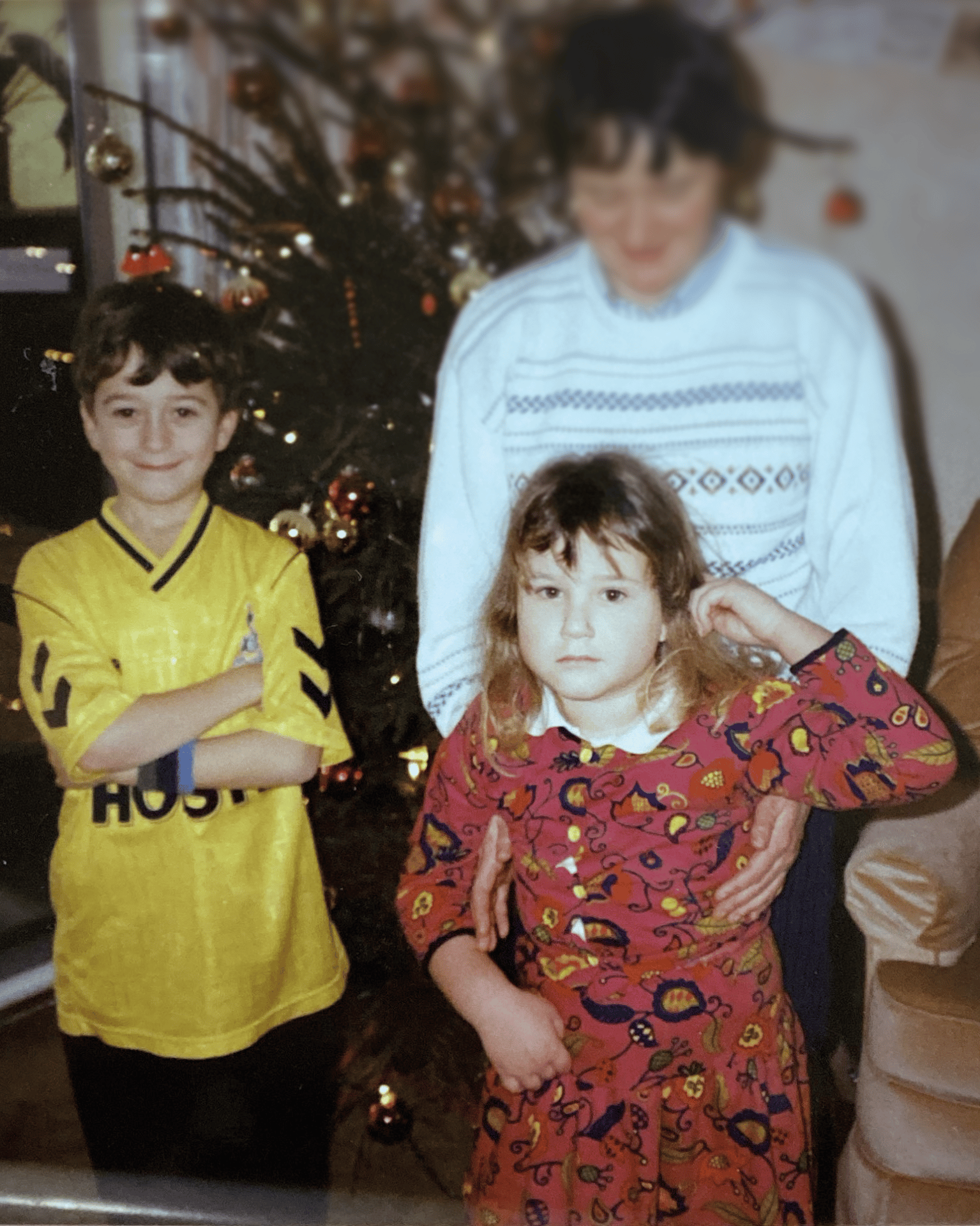 Two children, a boy in yellow and a girl in red, pose in front of a Christmas tree.
