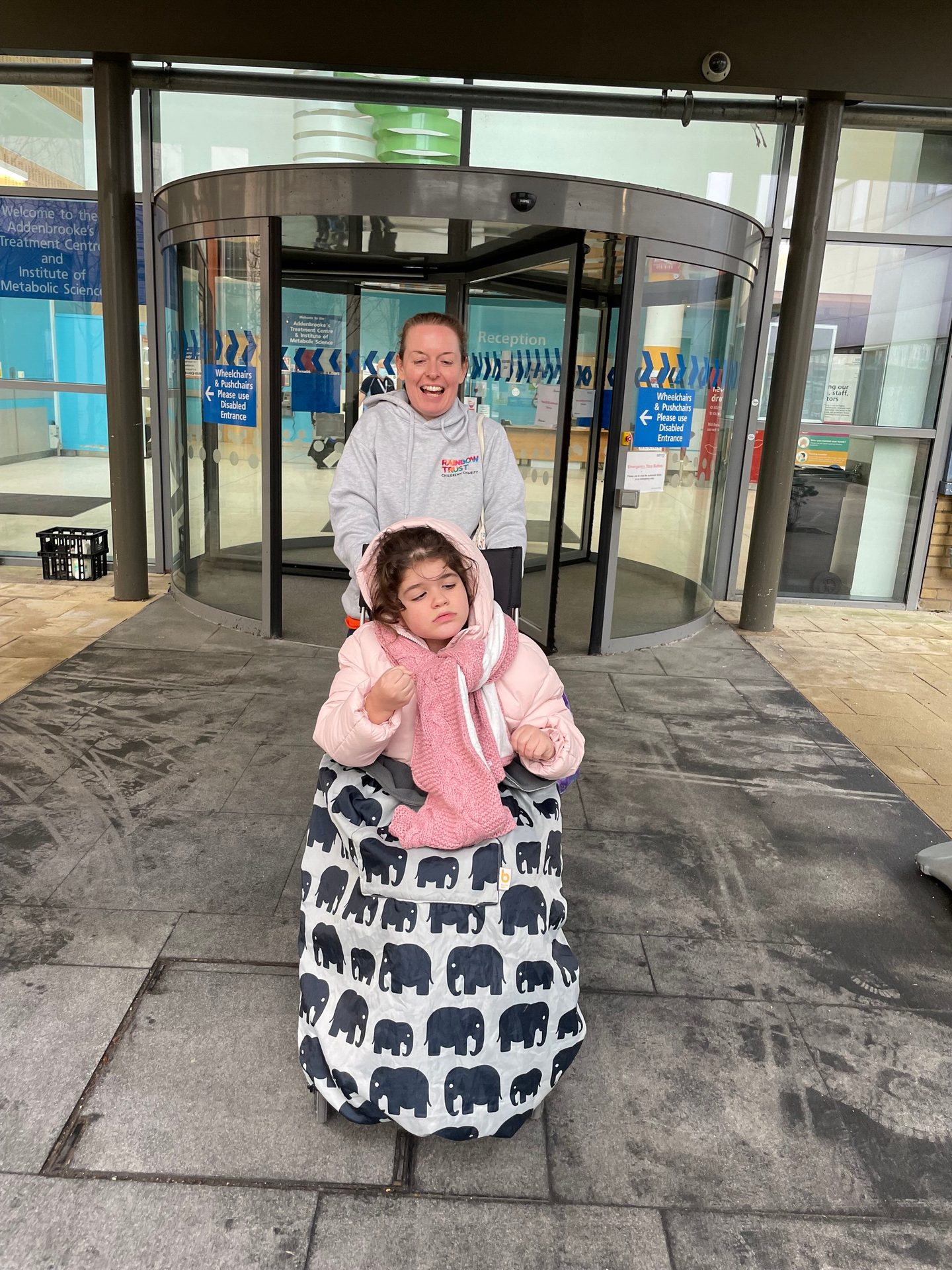 Smiling woman pushes child in wheelchair from Addenbrooke's hospital entrance.