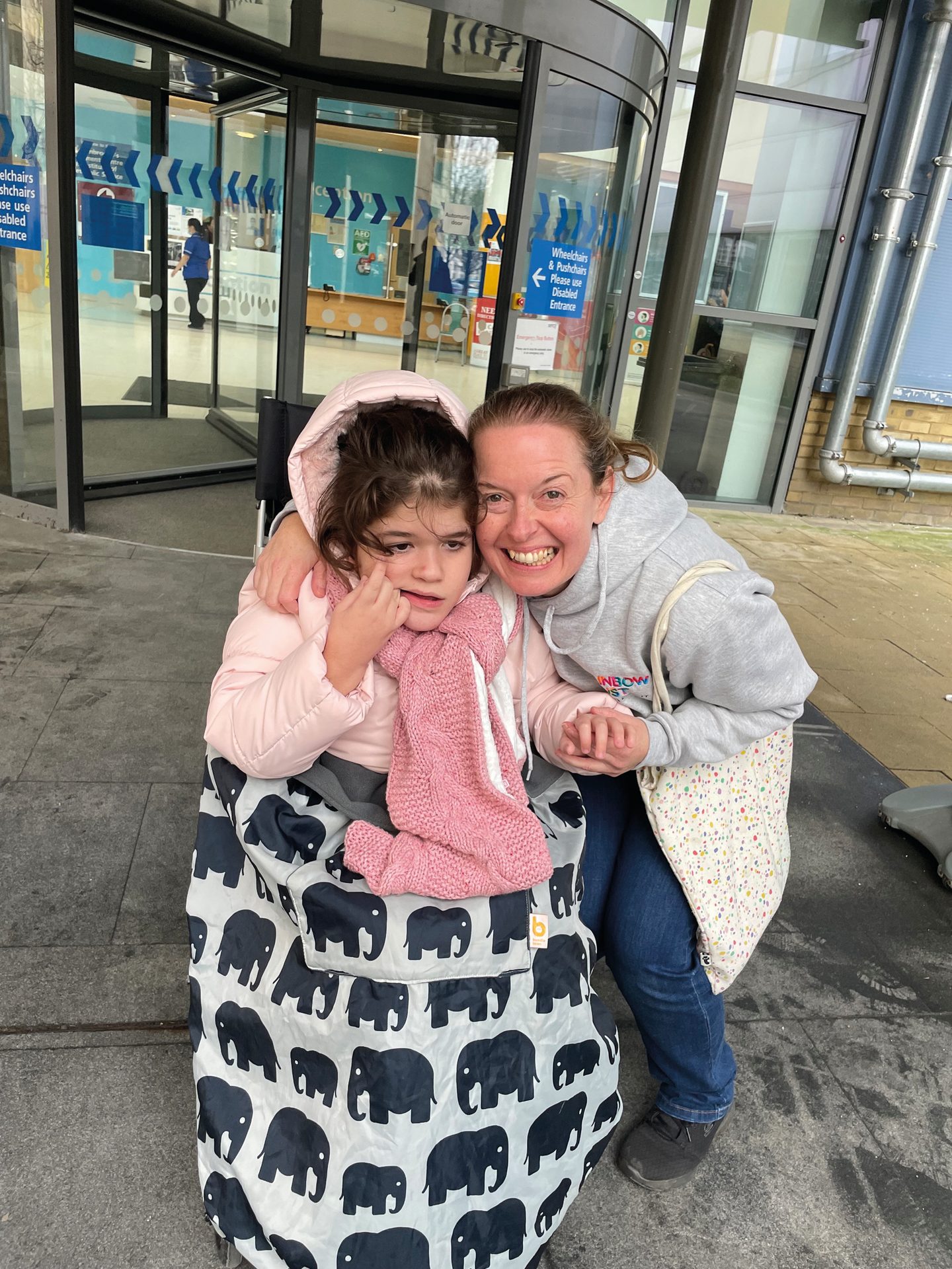 Woman embraces smiling child in a wheelchair outside a building entrance.